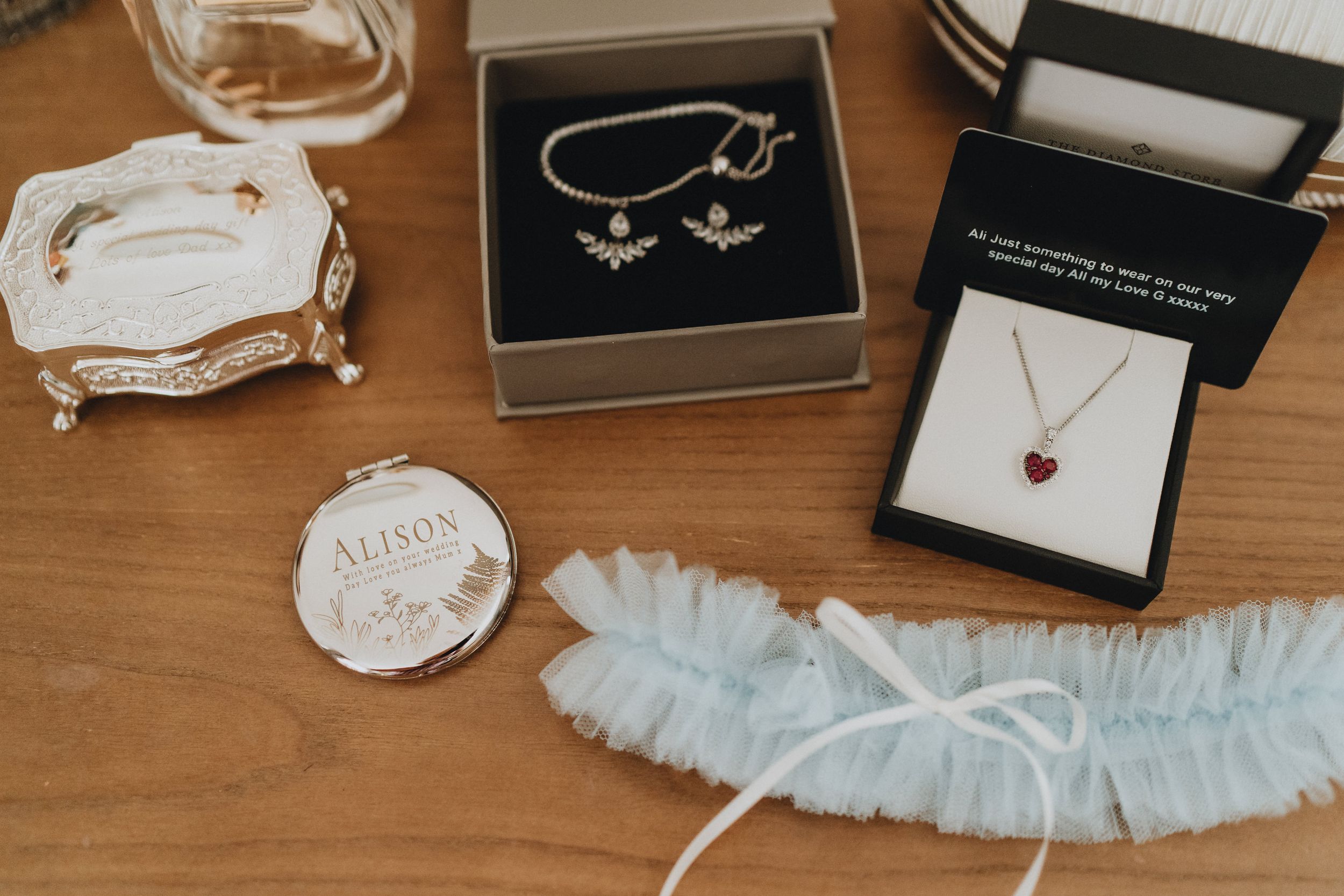 Delicate wedding accessories including jewelry boxes, a heart necklace, bridal garter, and name tag displayed on wooden surface.