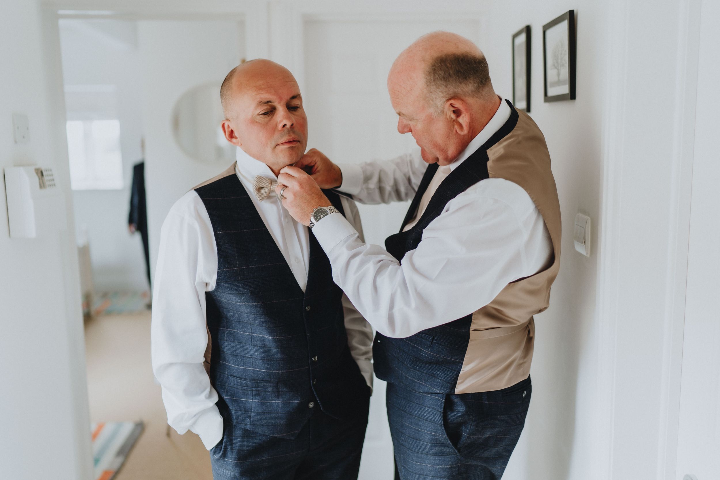 Two well-dressed men in waistcoats prepare for a formal event with one adjusting the other's bowtie.