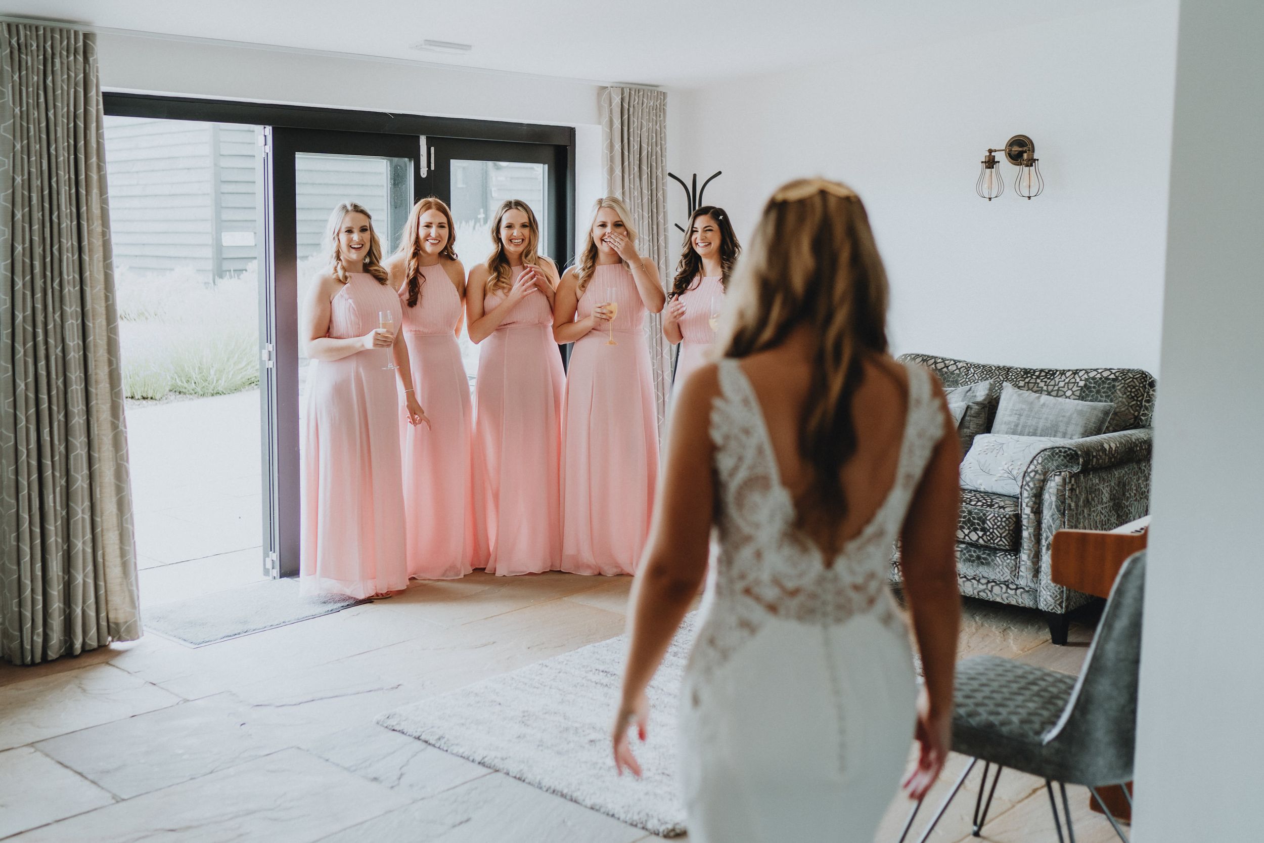 Bridesmaids in pink floor-length dresses react with delight as they look at someone in a white lace wedding gown.