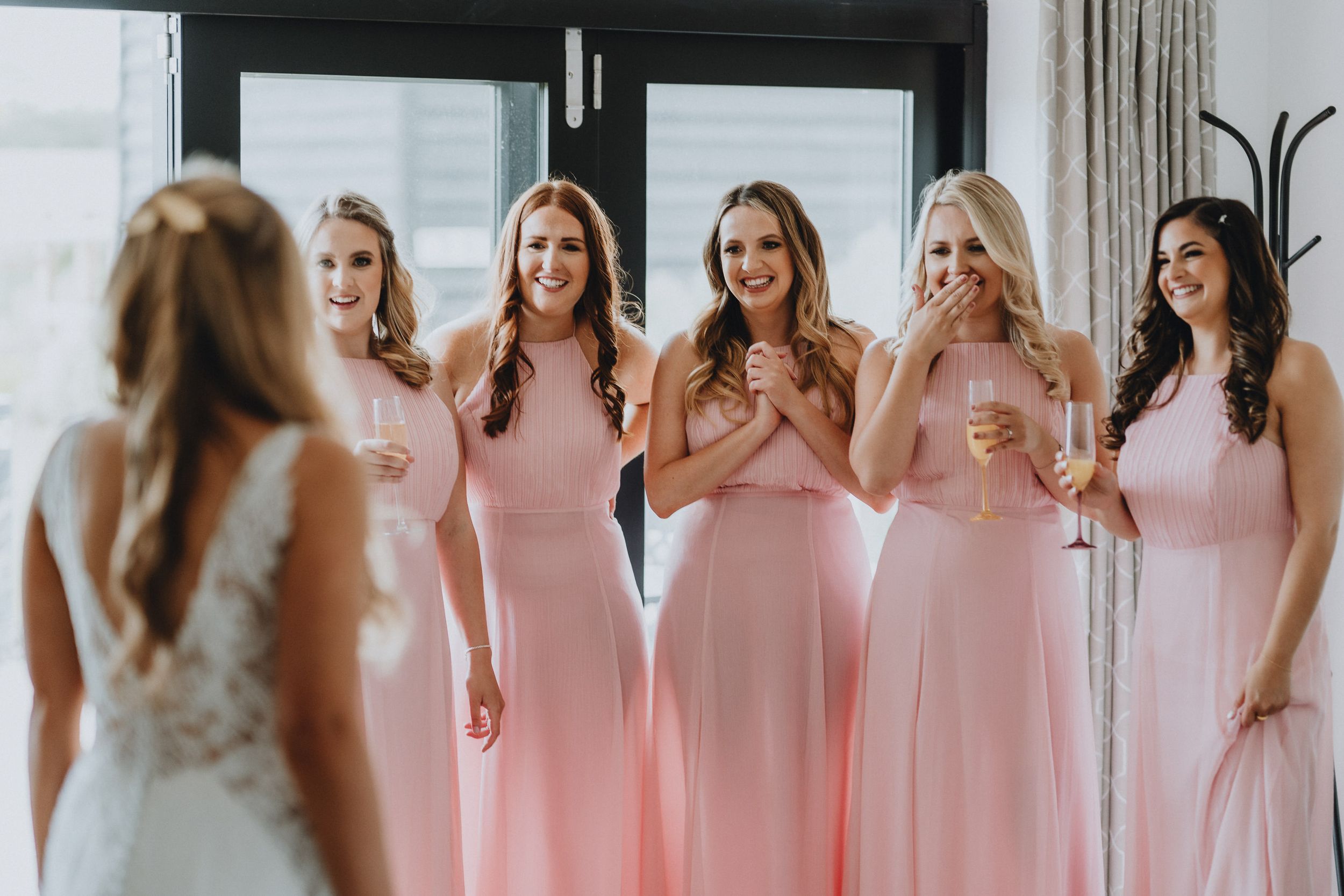 Bridesmaids in flowing pink dresses share a joyful moment during a wedding preparation.