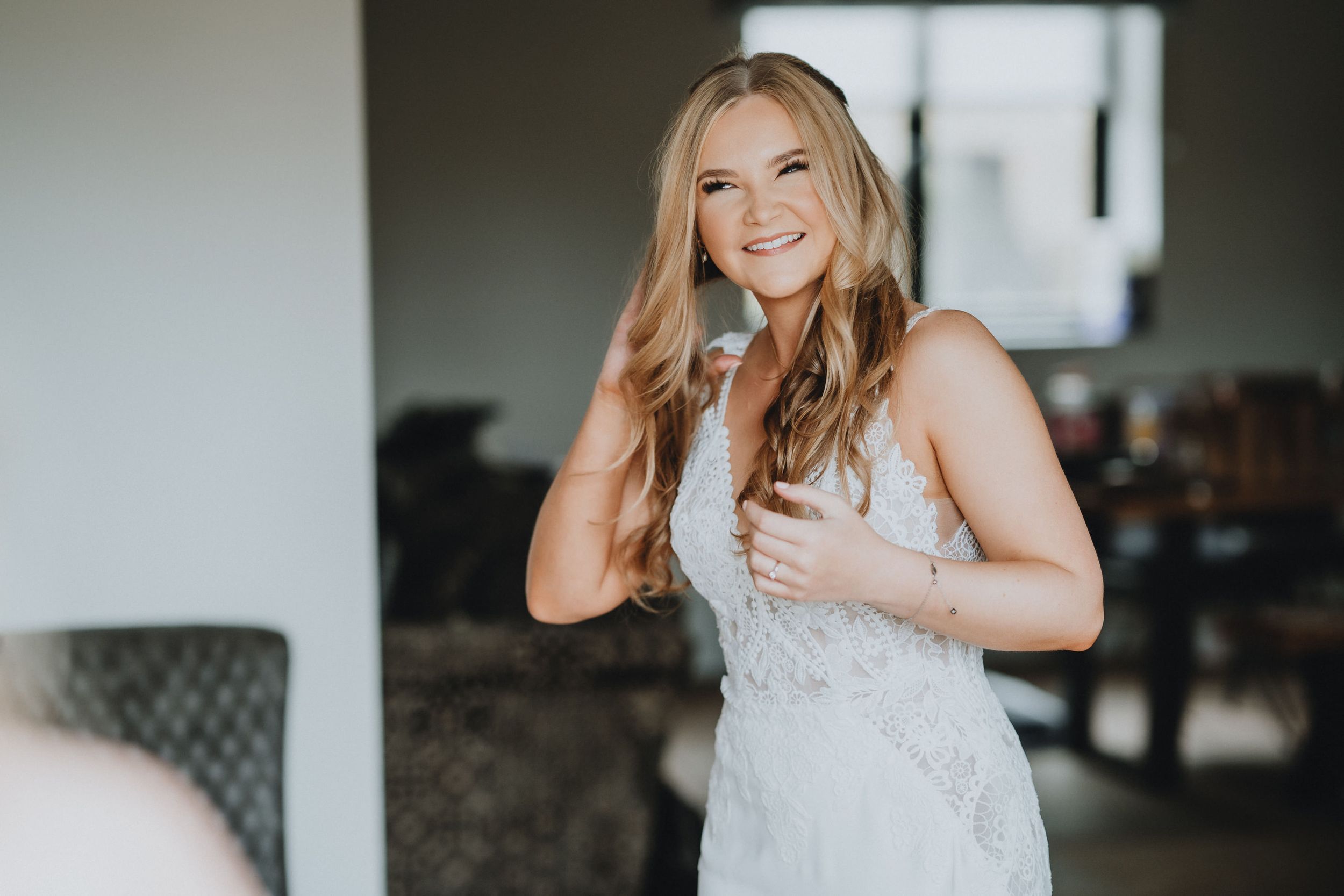 Smiling bride in white lace wedding dress stands indoors while touching her hair and holding a glass.