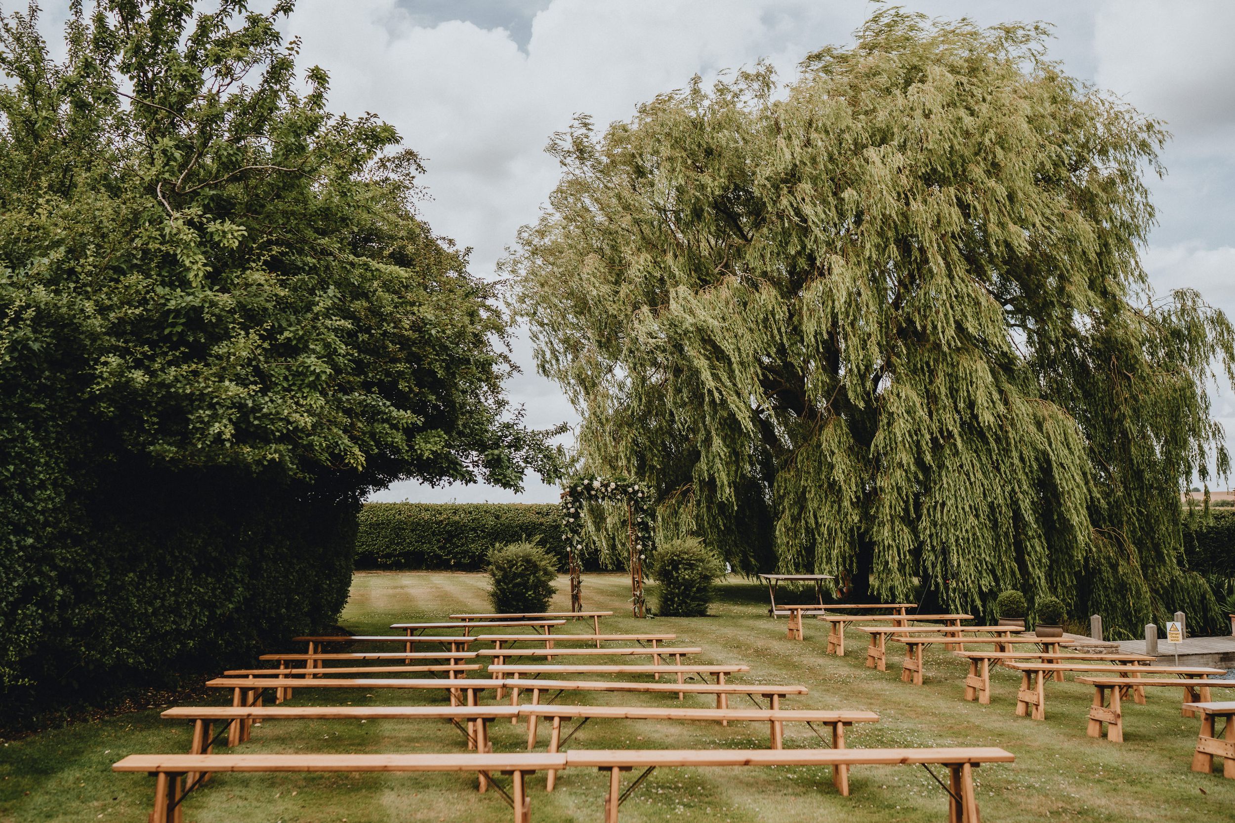 Wooden benches arranged in rows outdoors under a large weeping willow tree for an outdoor ceremony at Lattenbury Hill, Cambridgeshire.