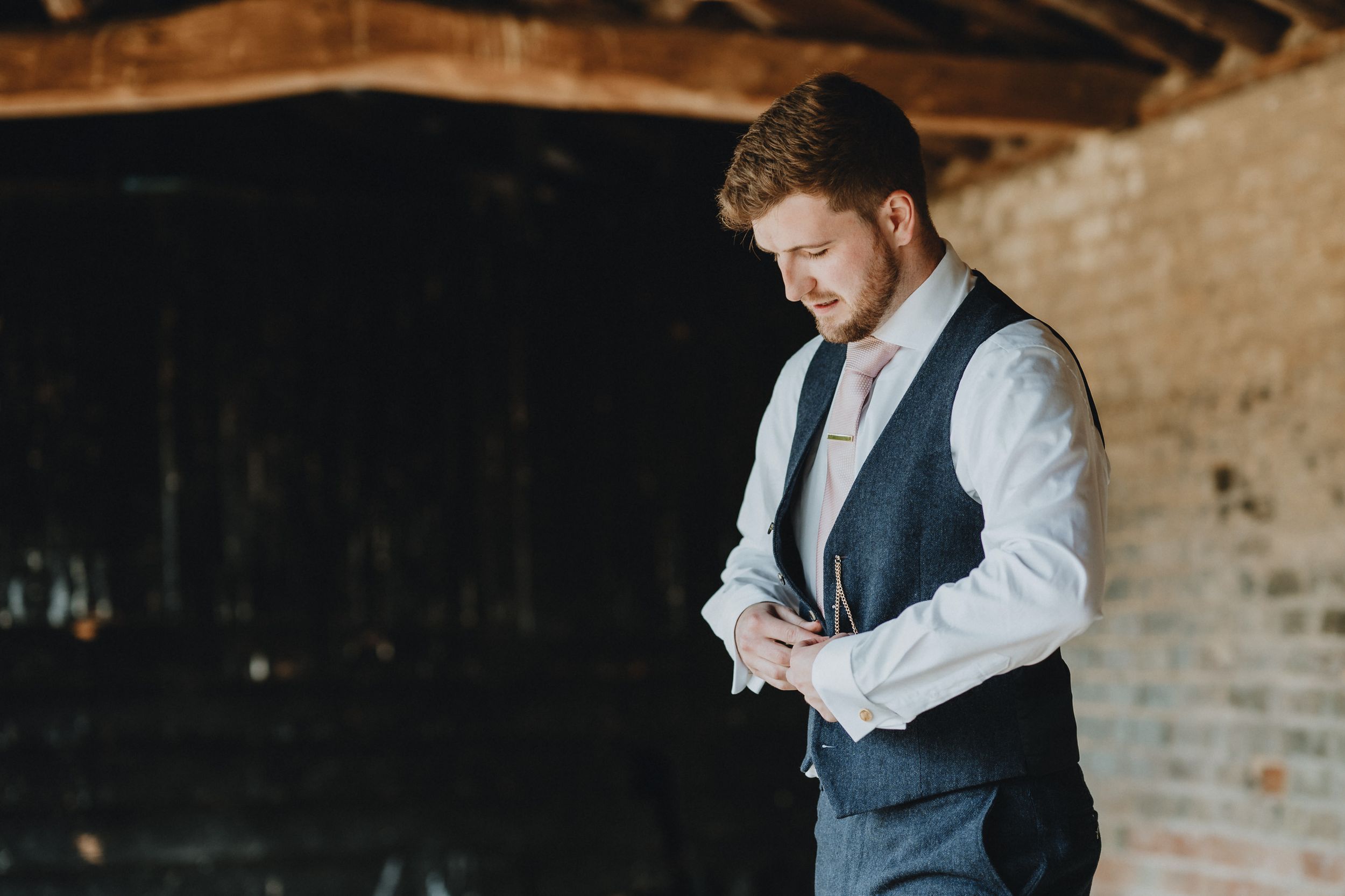 Man in formal vest and white shirt checks phone while standing against rustic brick wall in natural light.