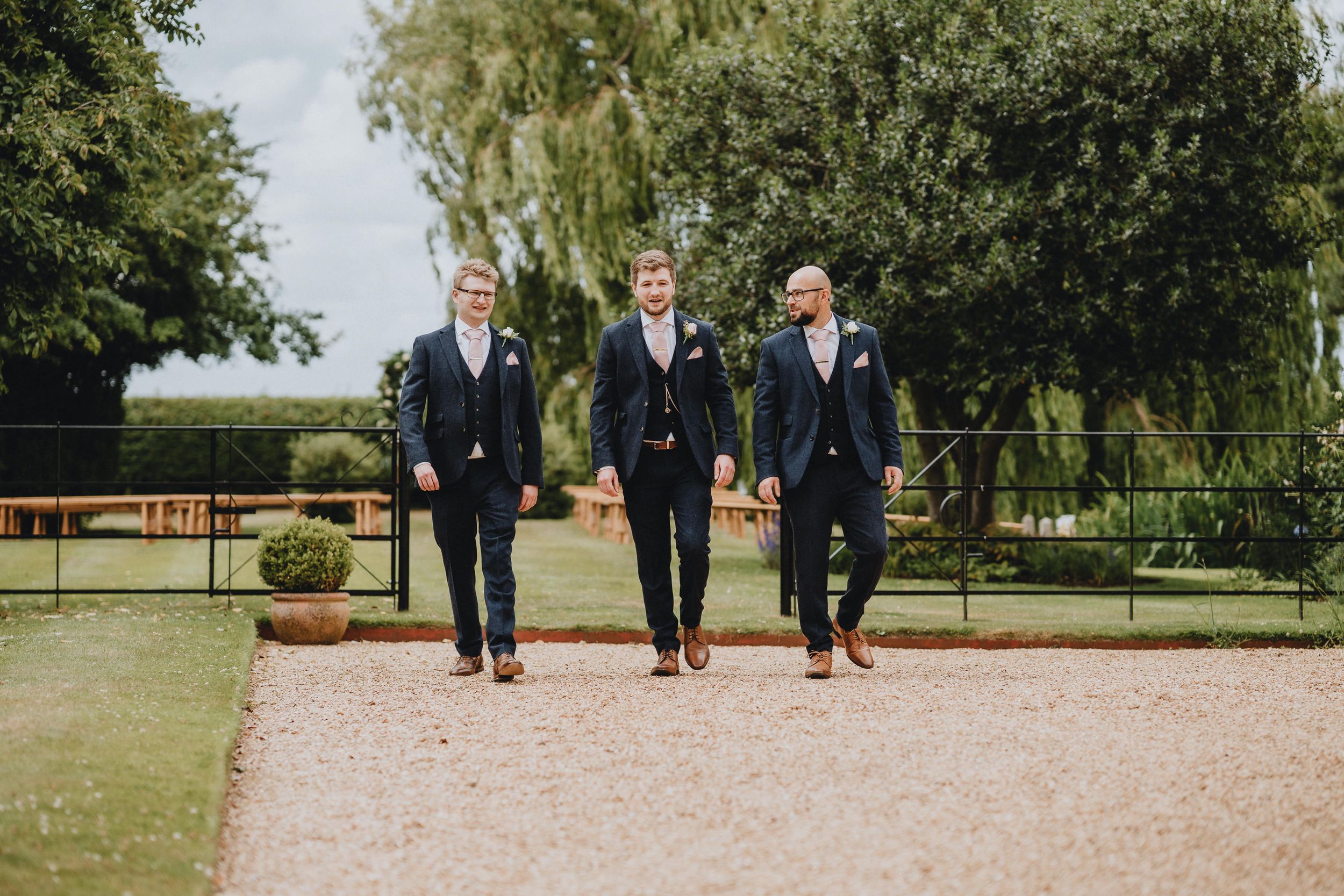 Three groomsmen in dark suits walk together on a gravel path in a garden setting.