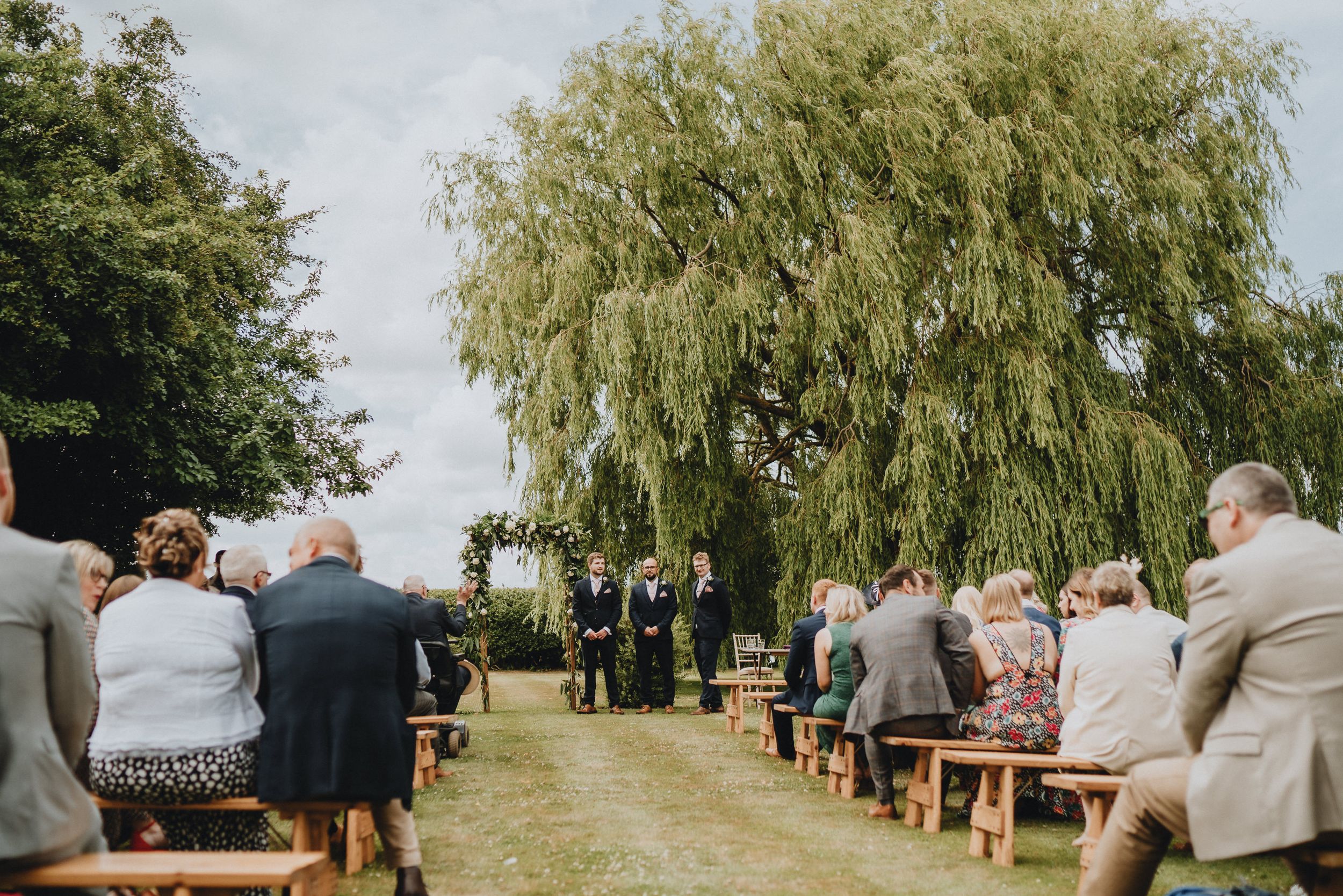 Wooden benches line a garden path under a weeping willow tree during an outdoor wedding ceremony.
