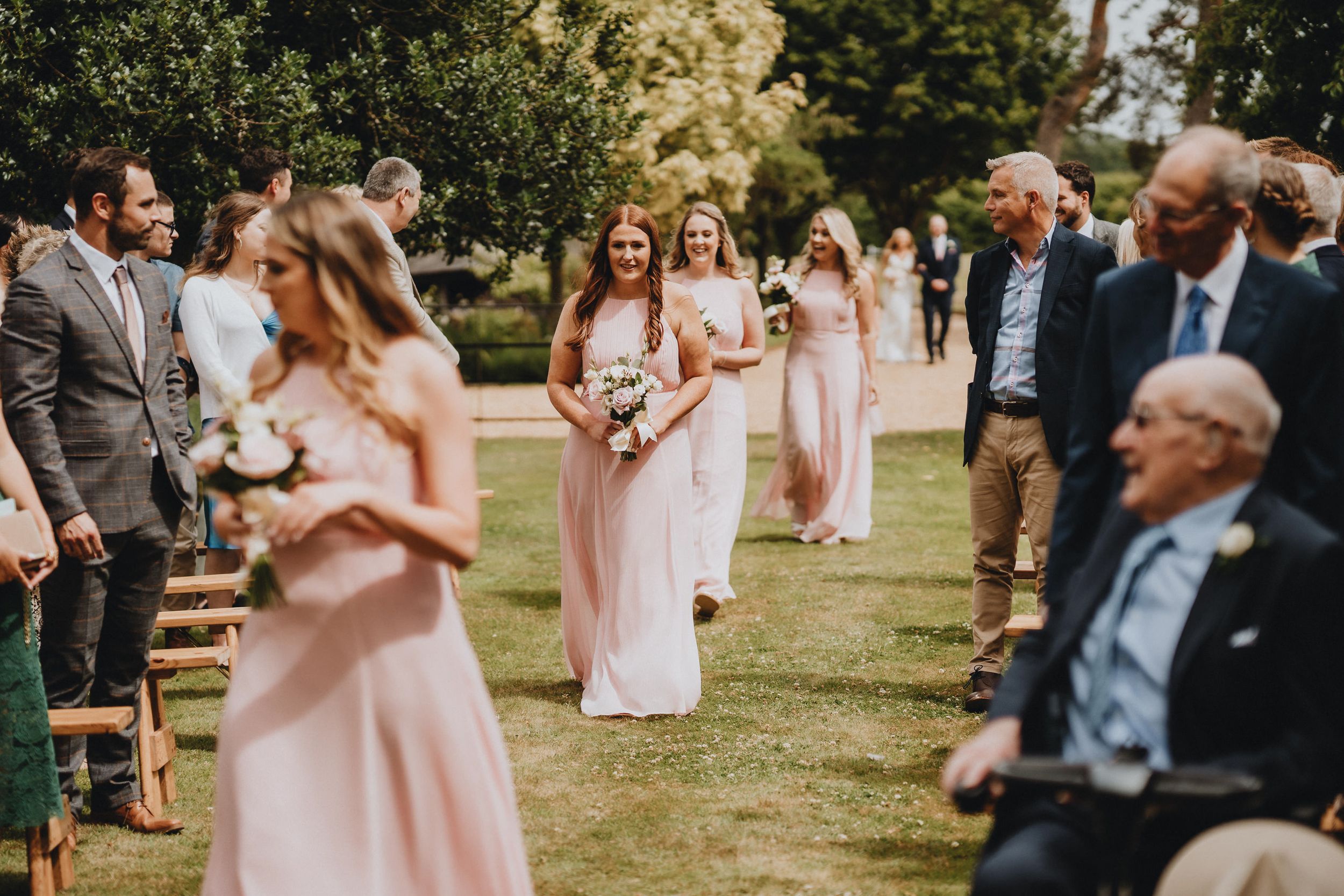 Wedding guests gather on a sunny lawn with people wearing pink dresses and gray suits during an outdoor celebration.