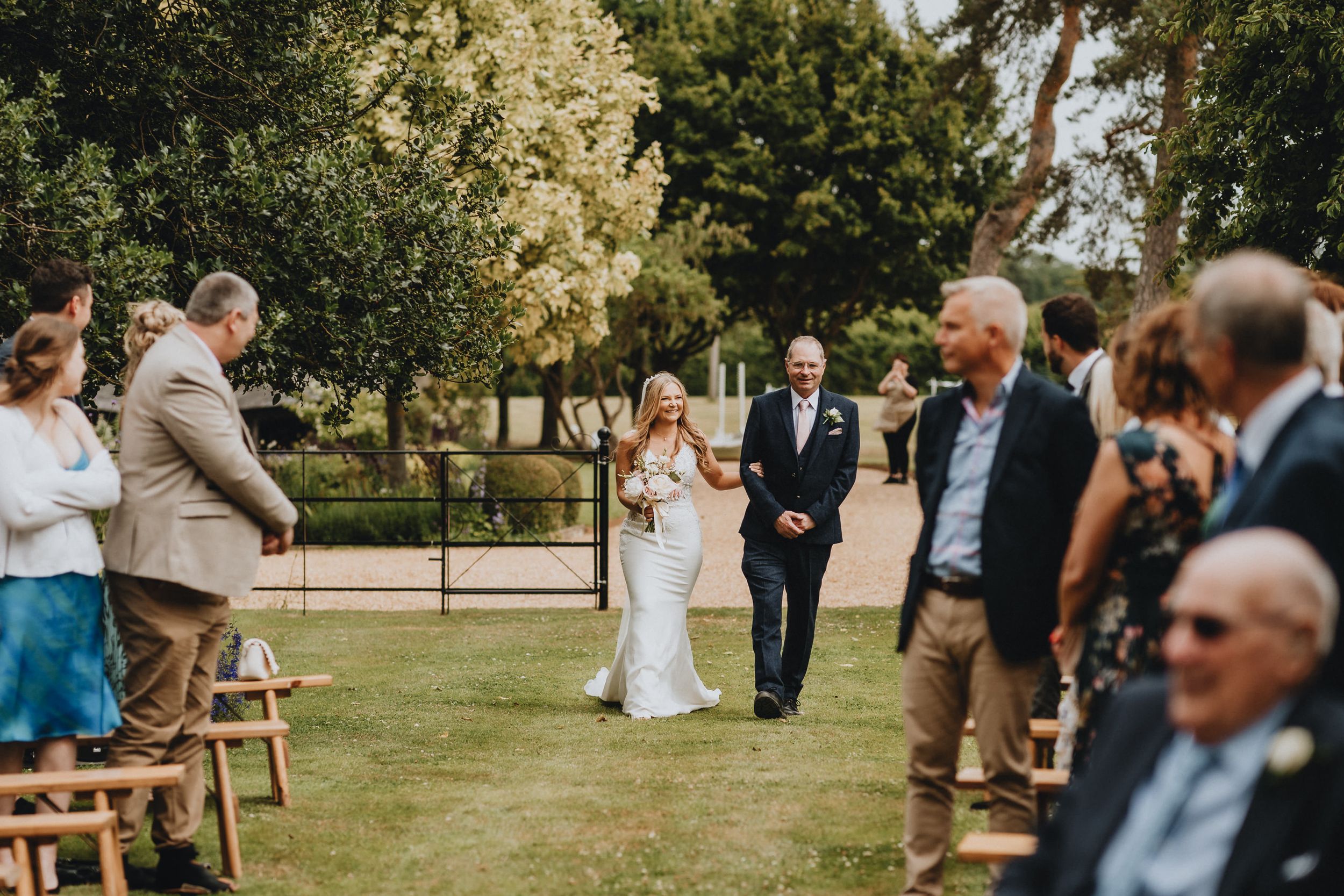 An outdoor wedding ceremony takes place in a lush garden setting with sunlit trees in the background.