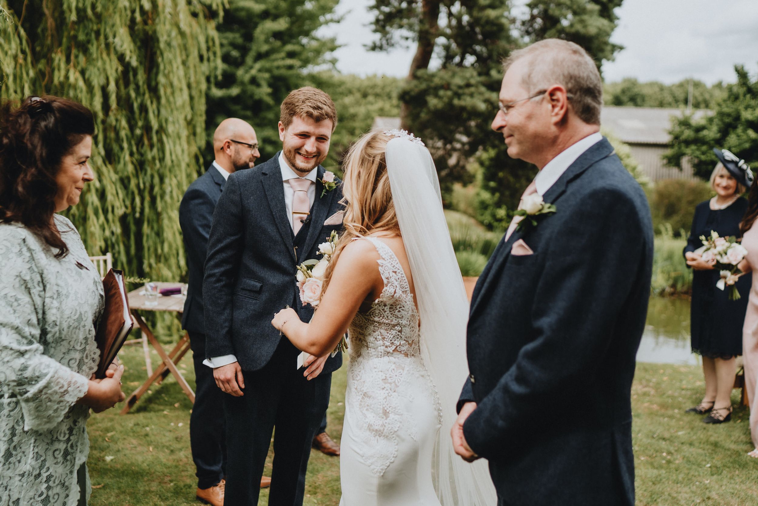 Outdoor wedding ceremony takes place by a willow tree beside a lake on a sunny summer day.