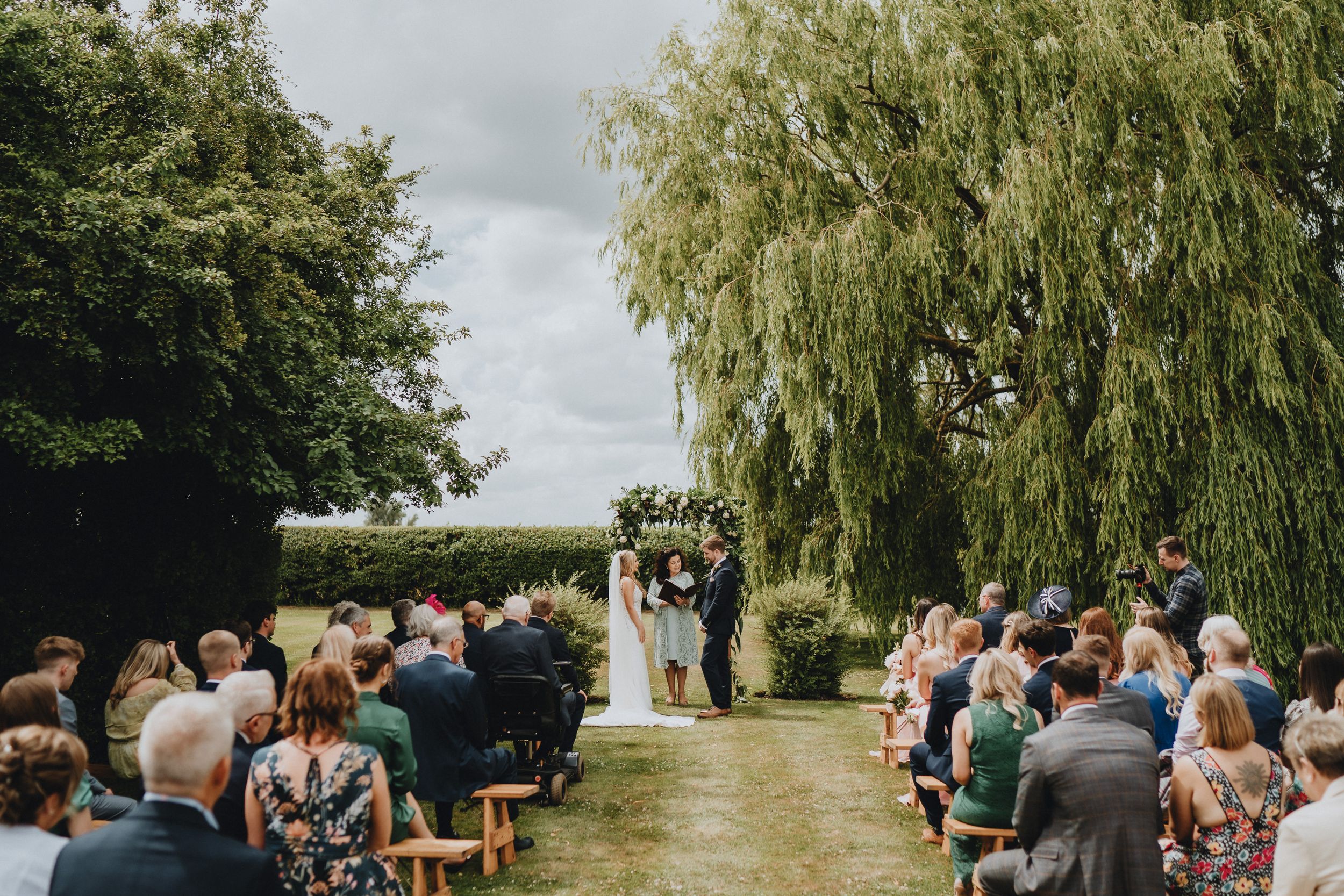 Outdoor garden wedding ceremony with guests seated in rows beneath a weeping willow tree on a cloudy day.