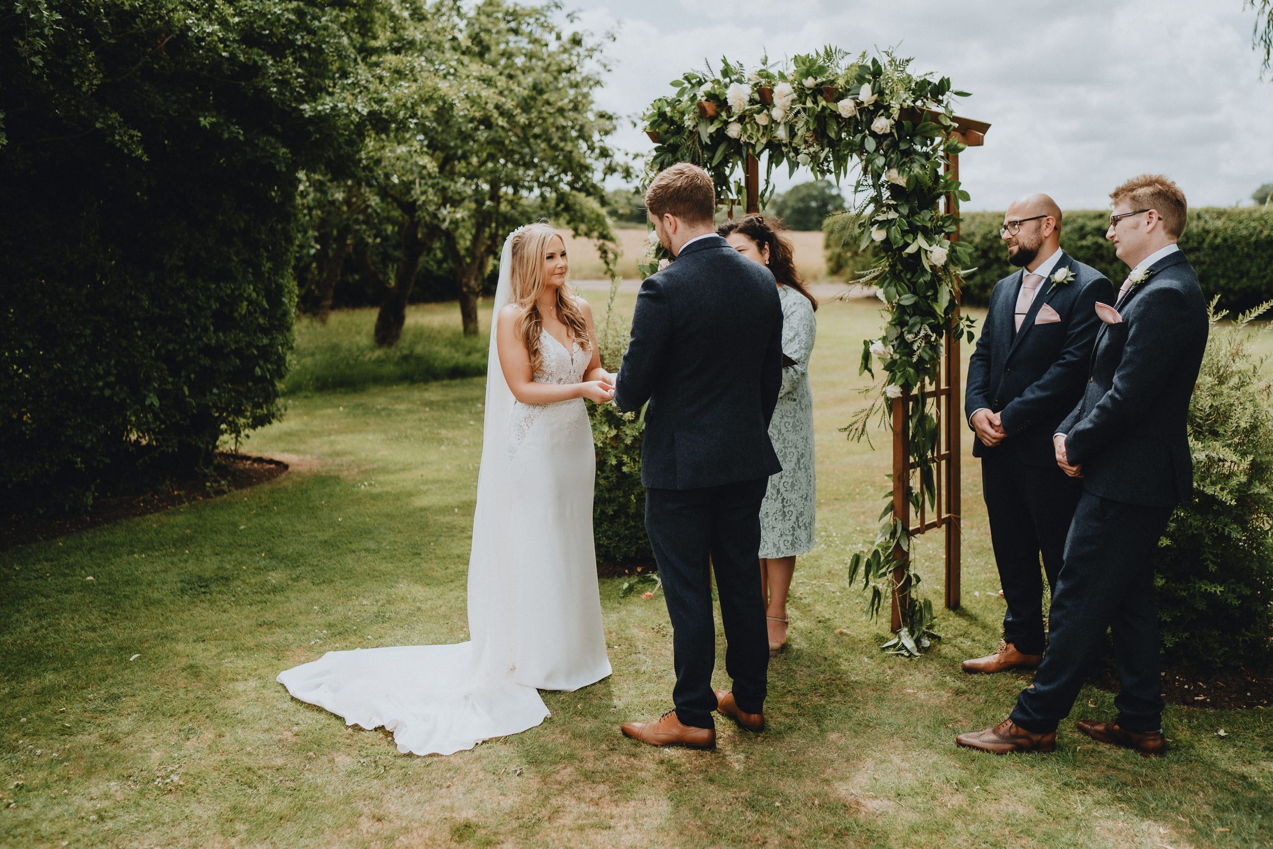 An intimate outdoor wedding ceremony takes place under a wooden arch adorned with greenery on a cloudy summer day.