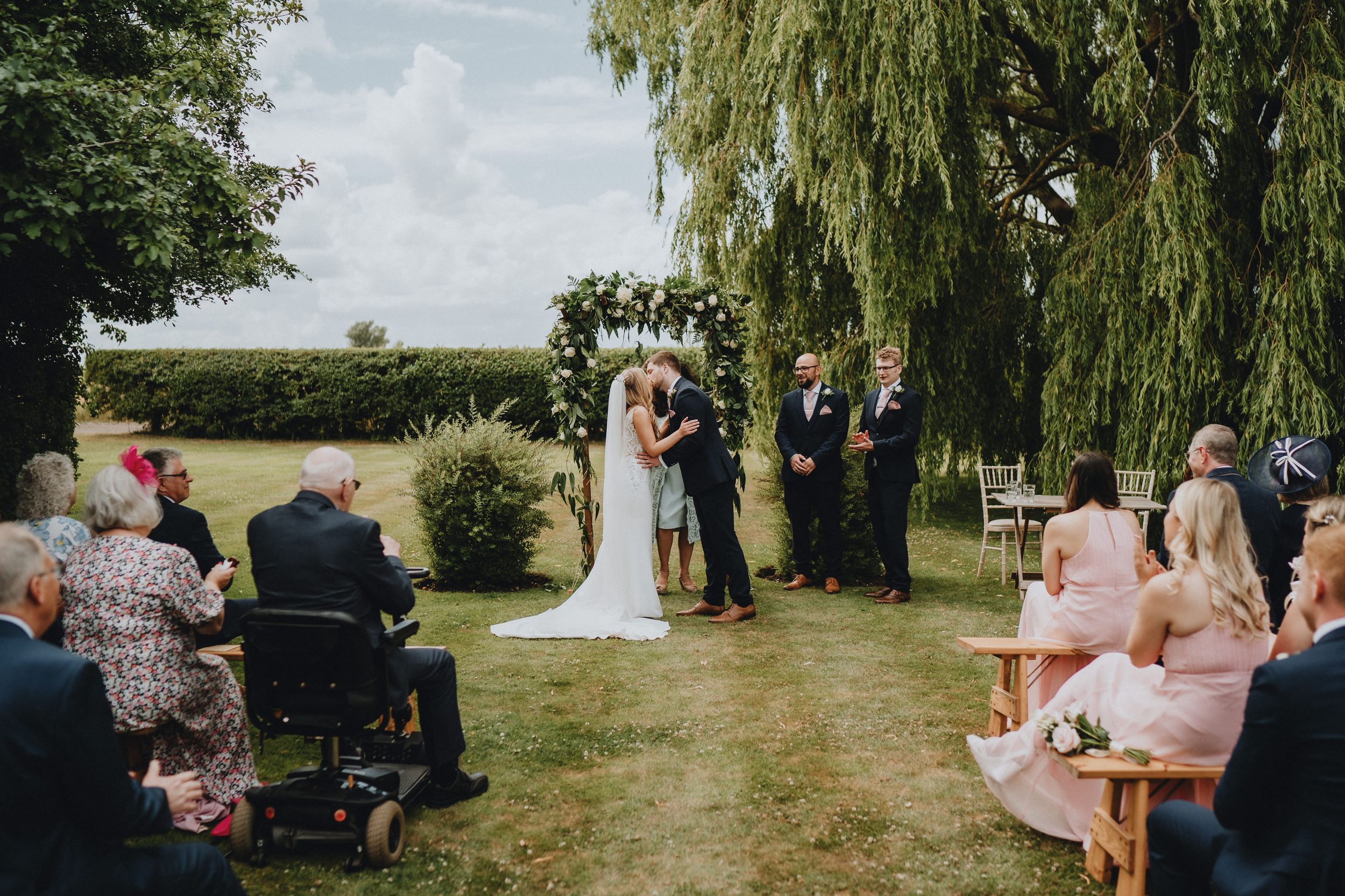 A beautiful outdoor wedding ceremony takes place under a willow tree with guests seated on a grassy lawn during a sunny day.
