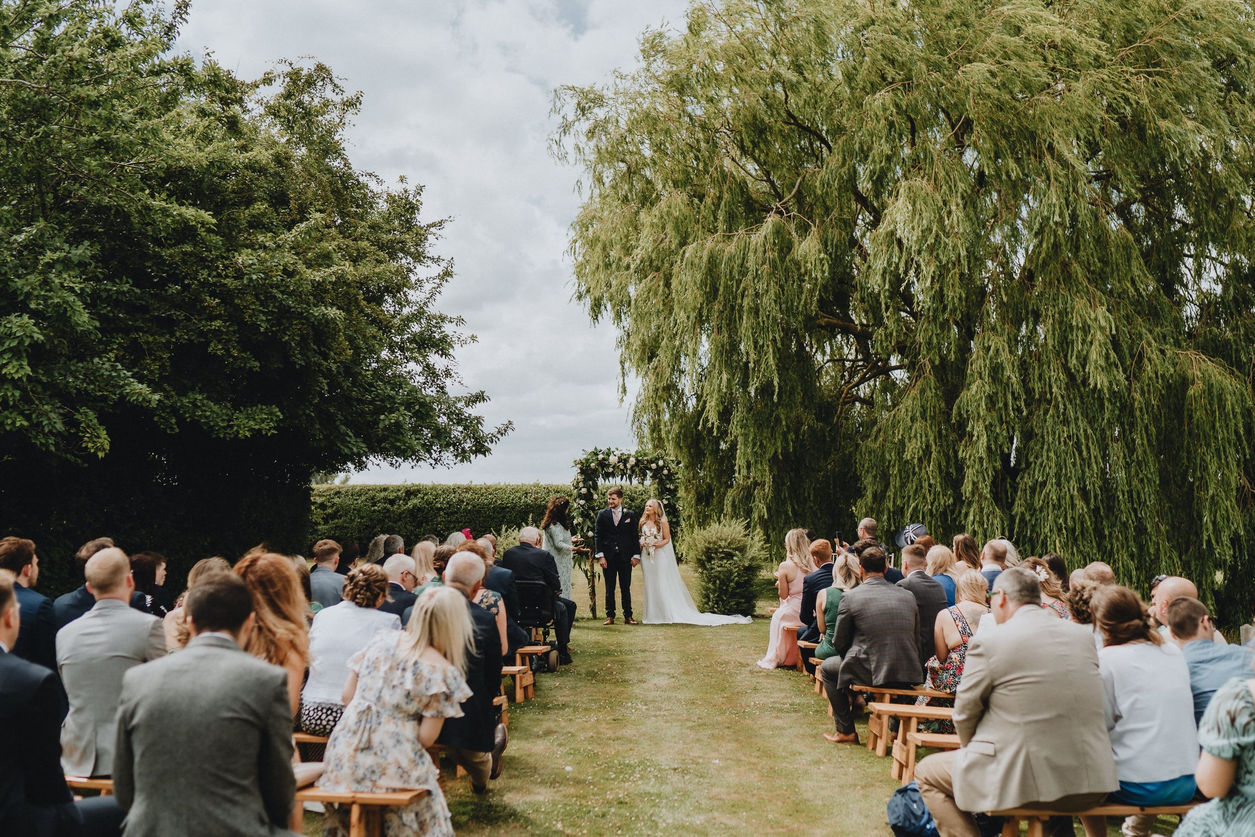 Outdoor wedding ceremony at Lattenbury Hill under weeping willows with guests seated on wooden chairs along a grassy aisle.