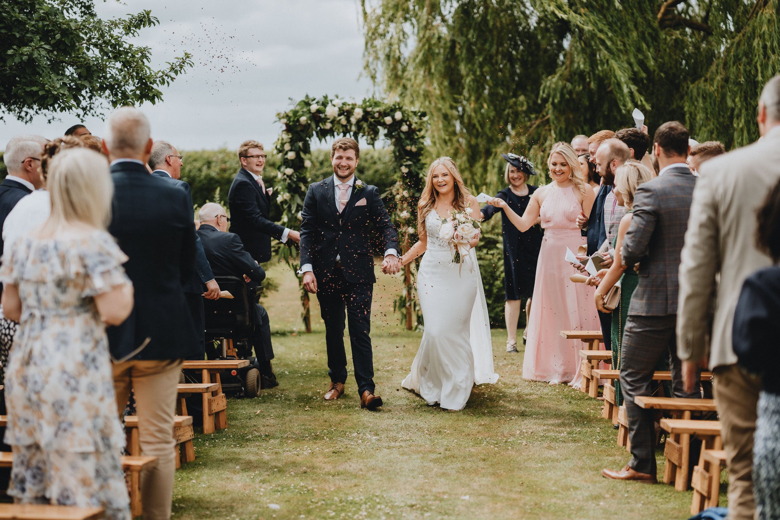 A joyful outdoor wedding ceremony under leafy trees with rustic wooden bench seating and floral arch decorations.
