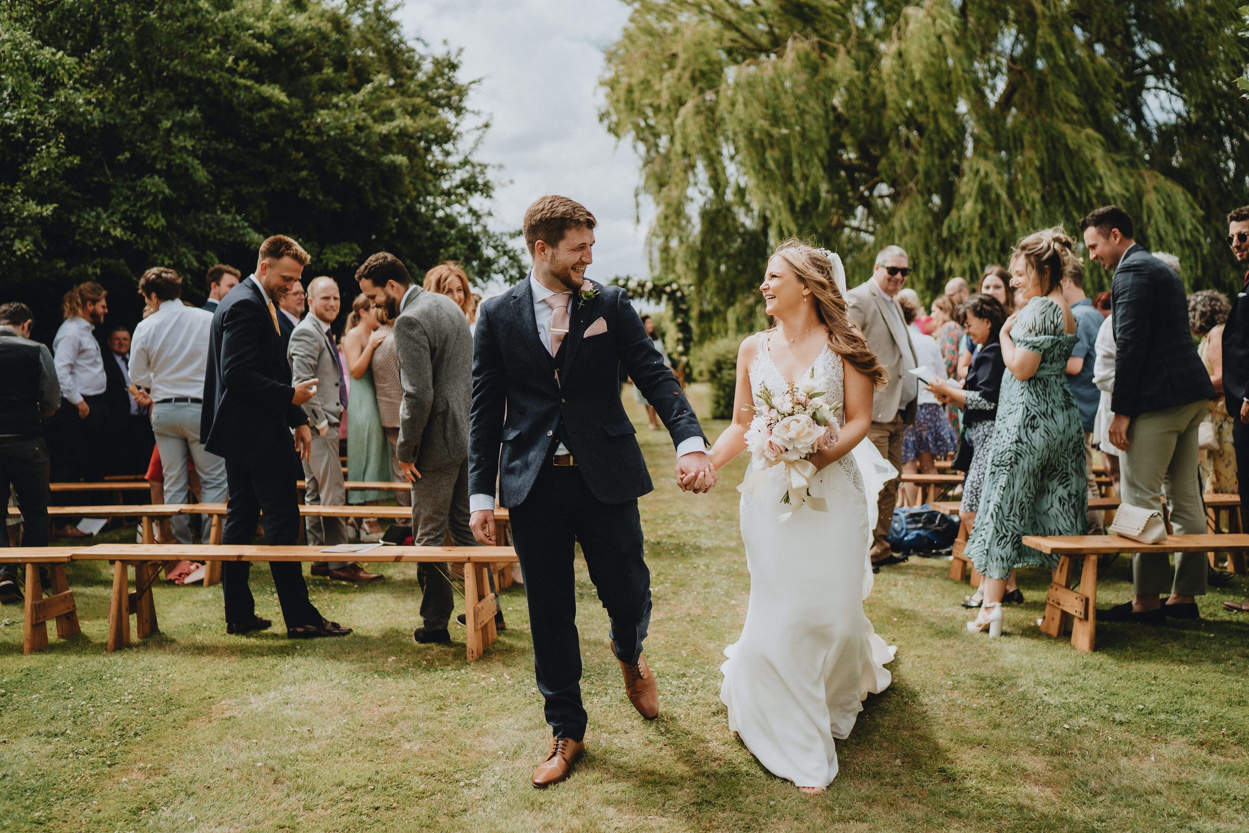 A couple celebrates at an outdoor wedding reception with wooden benches and guests gathered in a garden setting.