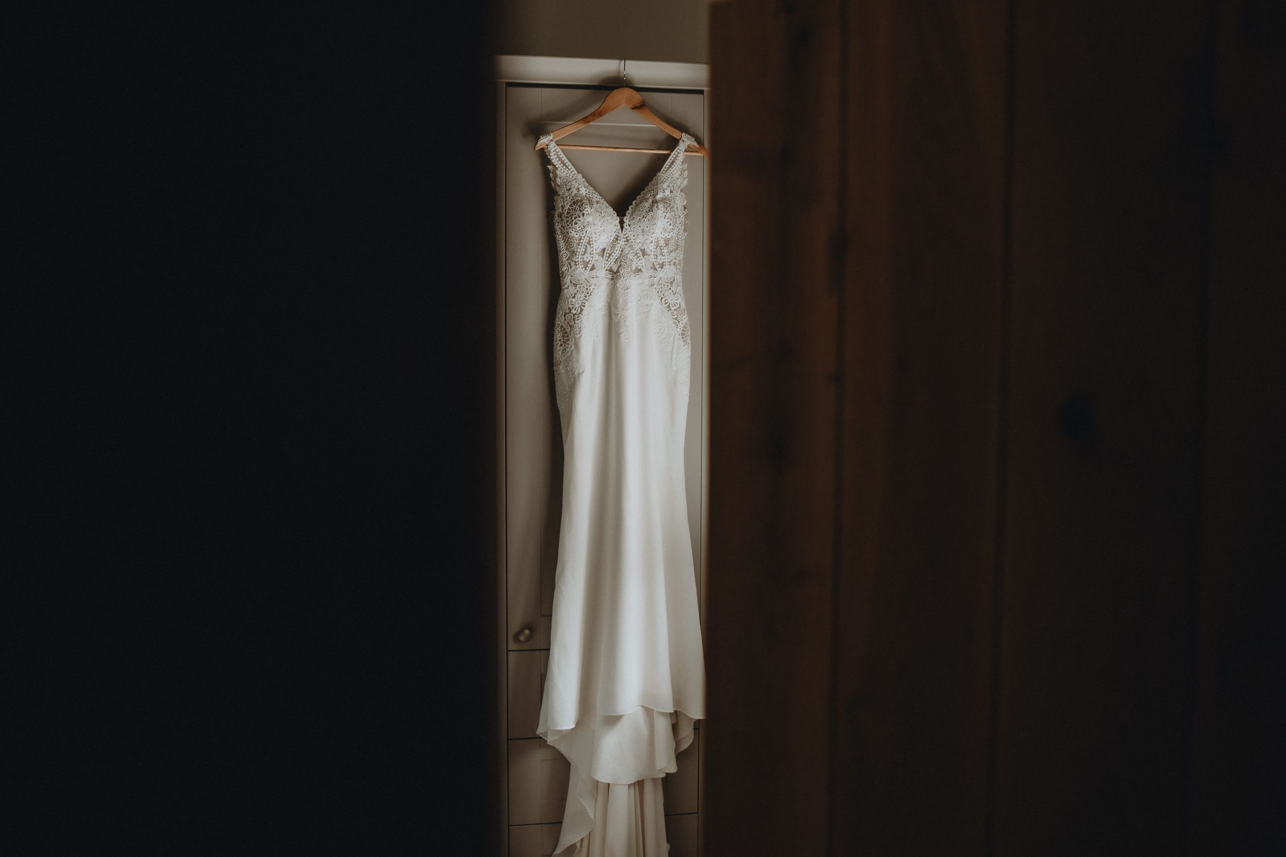 A white wedding dress with delicate lace details hangs elegantly against a dark brown wall in soft lighting.
