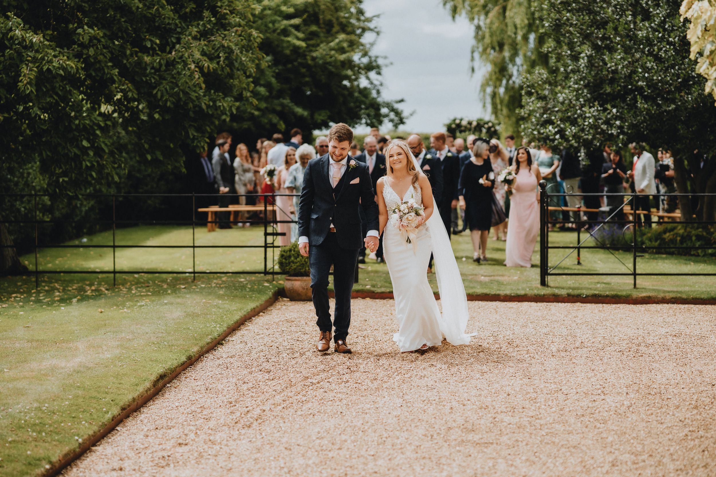 A couple walks down a gravel path at an outdoor wedding ceremony while guests look on from behind.