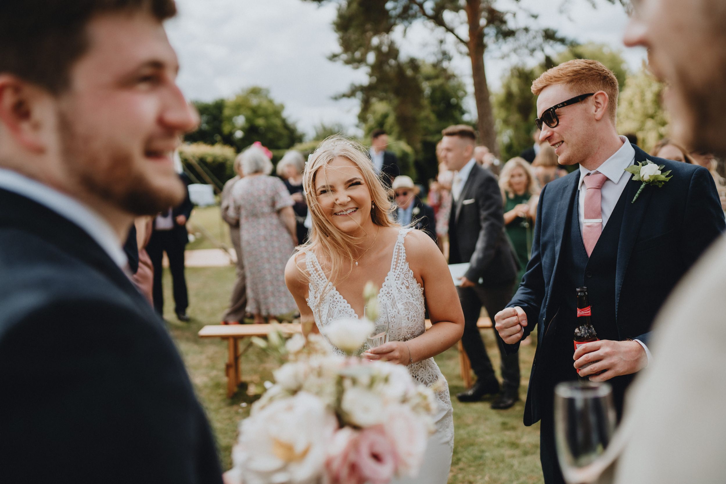A sunny outdoor wedding celebration with guests gathered on a green lawn, featuring elegant floral bouquets.