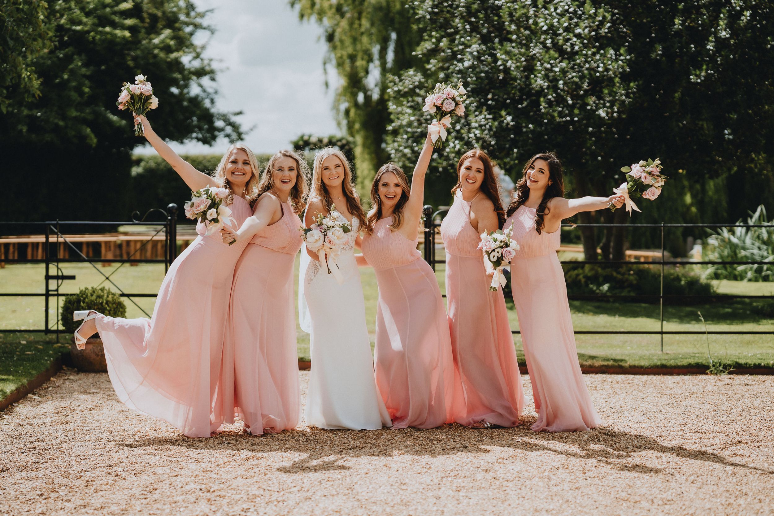A cheerful bridal party in flowing pink gowns poses together outdoors with bouquets raised in celebration.