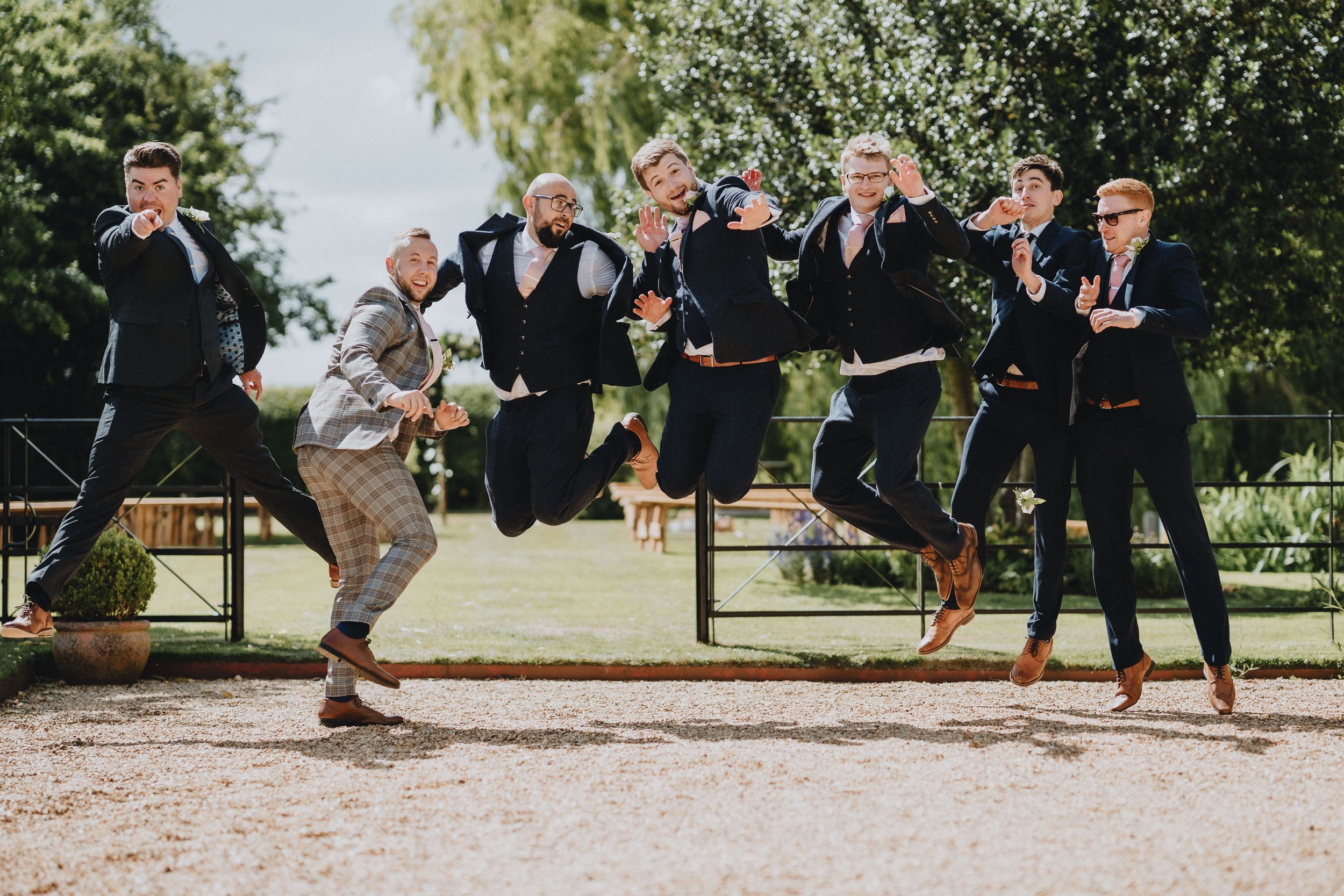 A joyful group in formal attire leaps in unison on a gravel path in a garden setting.