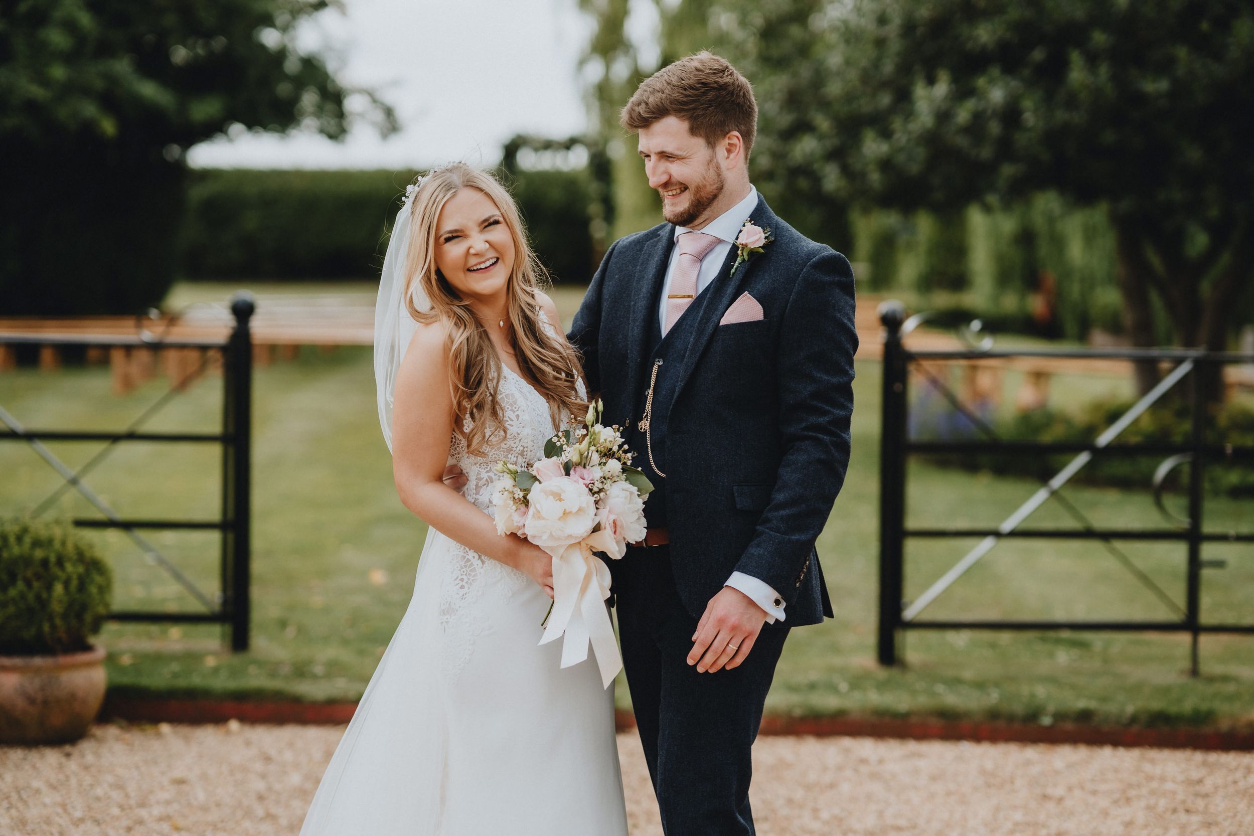 A couple poses together at their outdoor wedding with rustic wooden gates and greenery in the background.