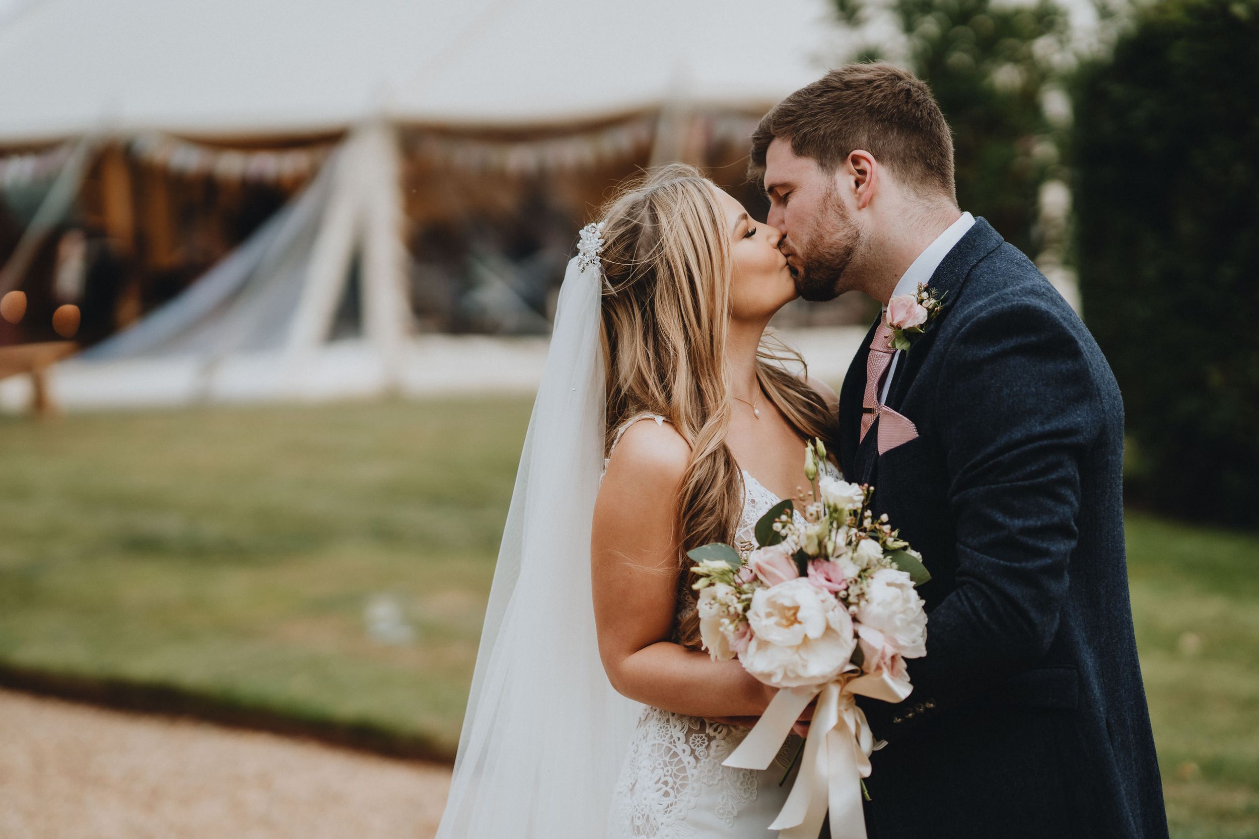 Romantic outdoor wedding scene with couple kissing by rustic tent and holding elegant white floral bouquet.