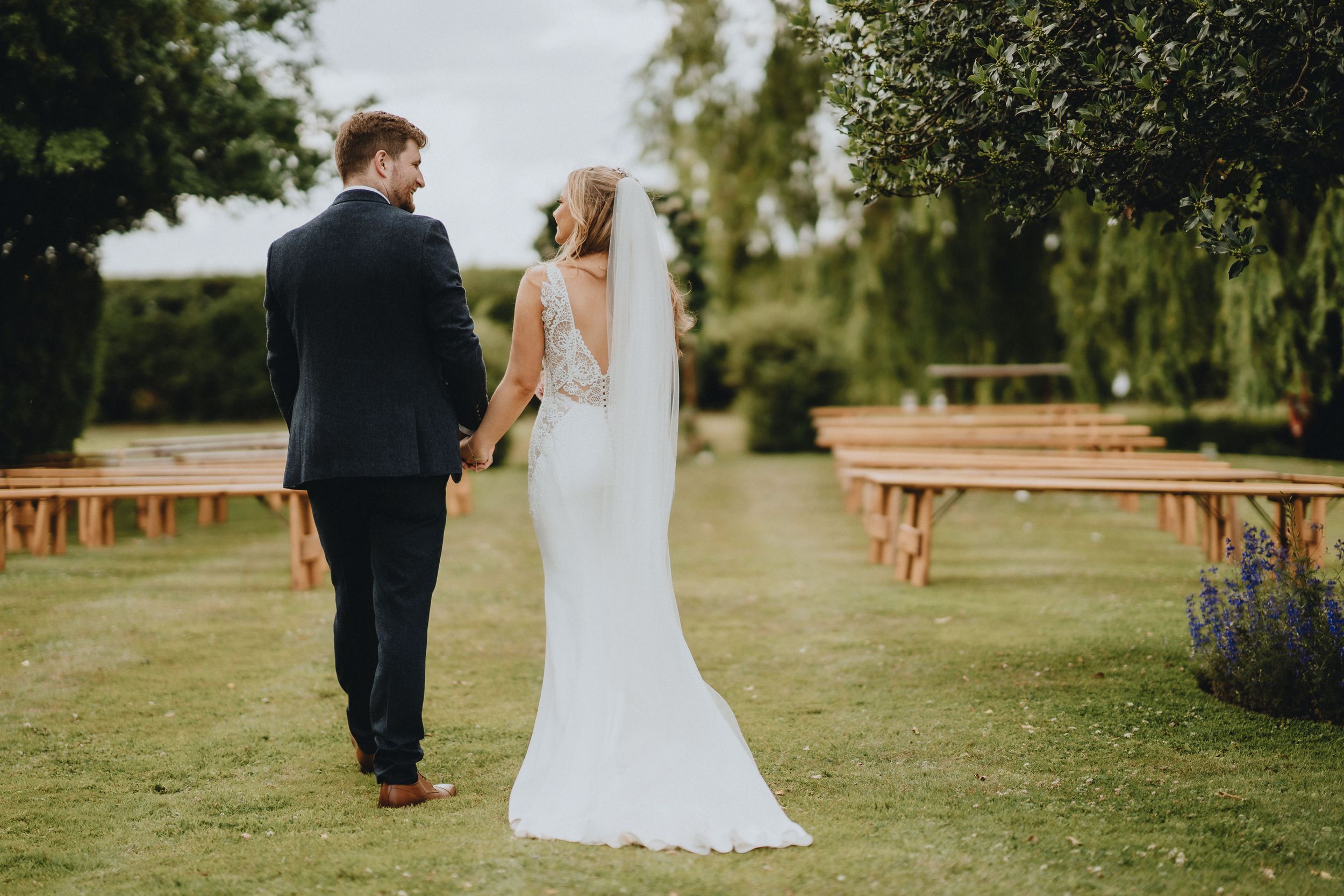 A newlywed couple holding hands while walking between wooden benches at an outdoor garden wedding ceremony.