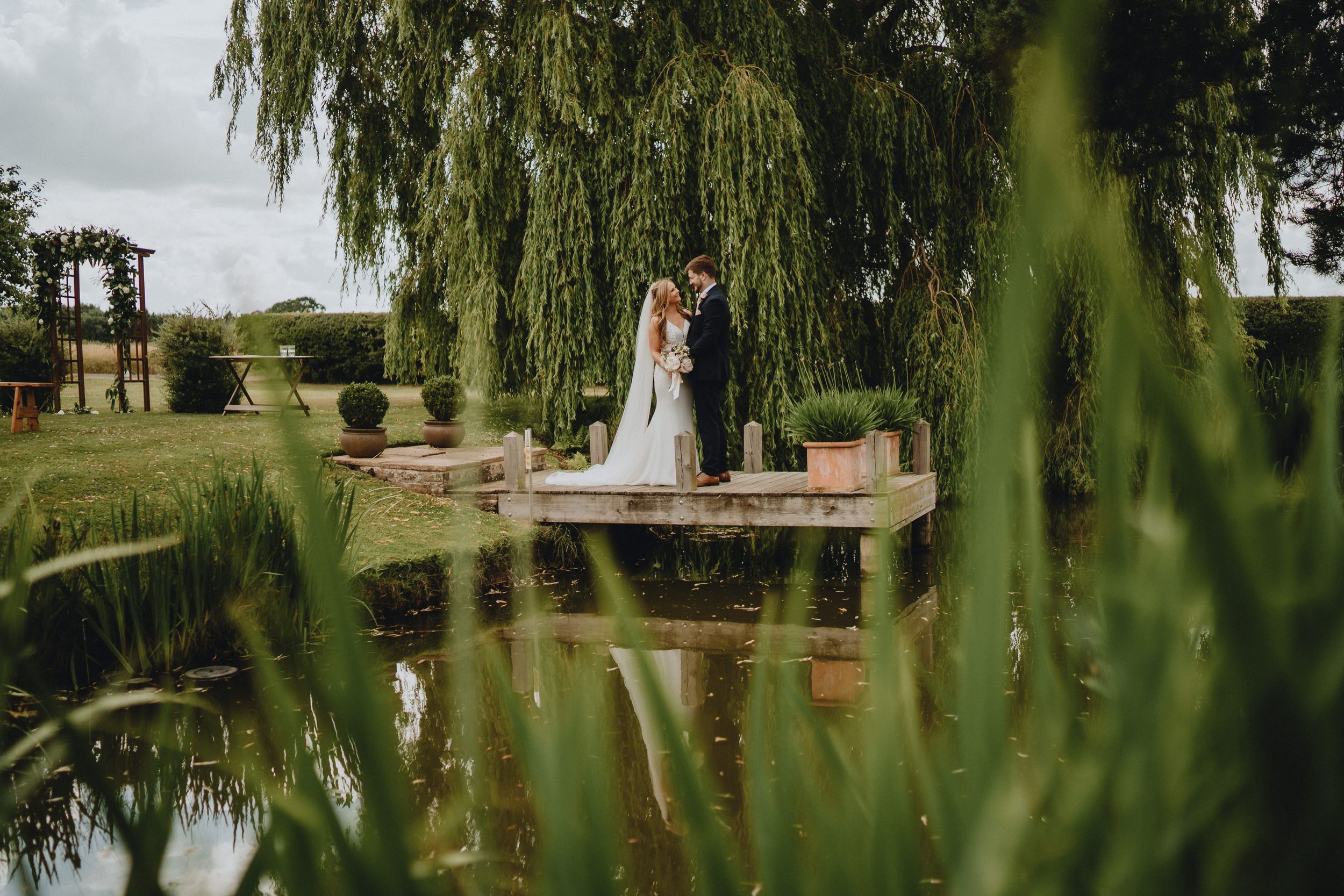 A couple in wedding attire stands together on a wooden pier overlooking a tranquil pond with a weeping willow tree.
