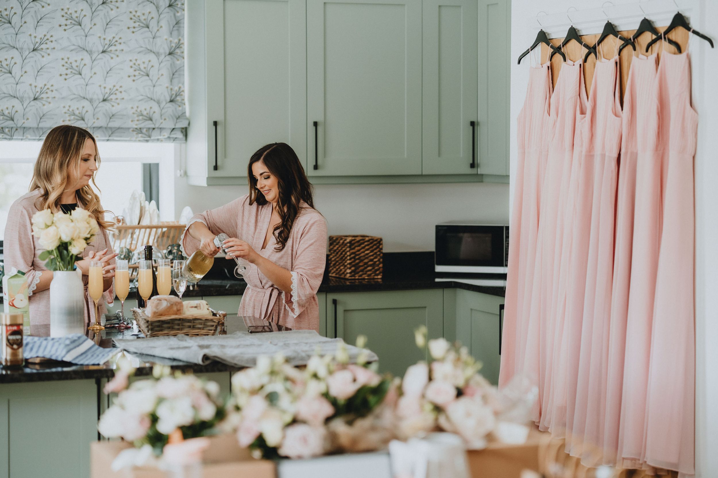 Wedding party members arrange flowers in a kitchen while pink bridesmaid dresses hang nearby.