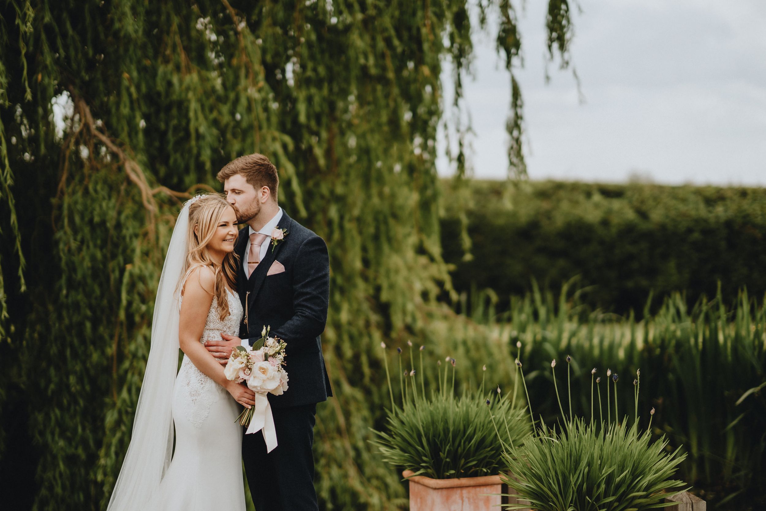 A couple embrace for a wedding photo under a weeping willow tree with a romantic countryside backdrop.
