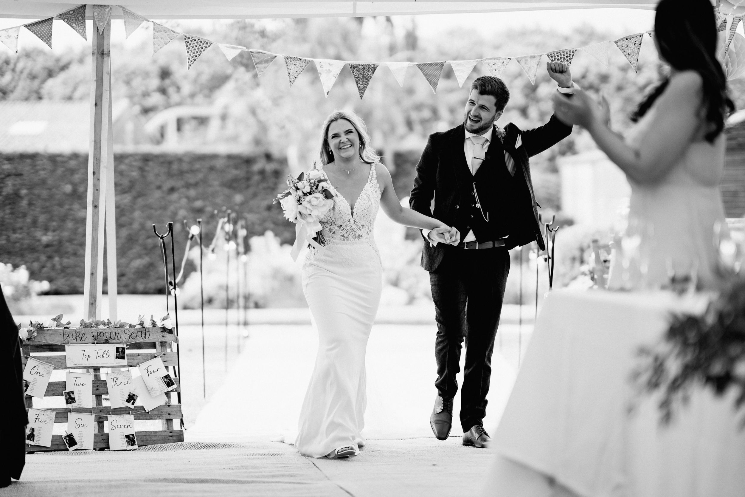Black and white photo of happy couple walking together at outdoor wedding reception under white tent with bunting.