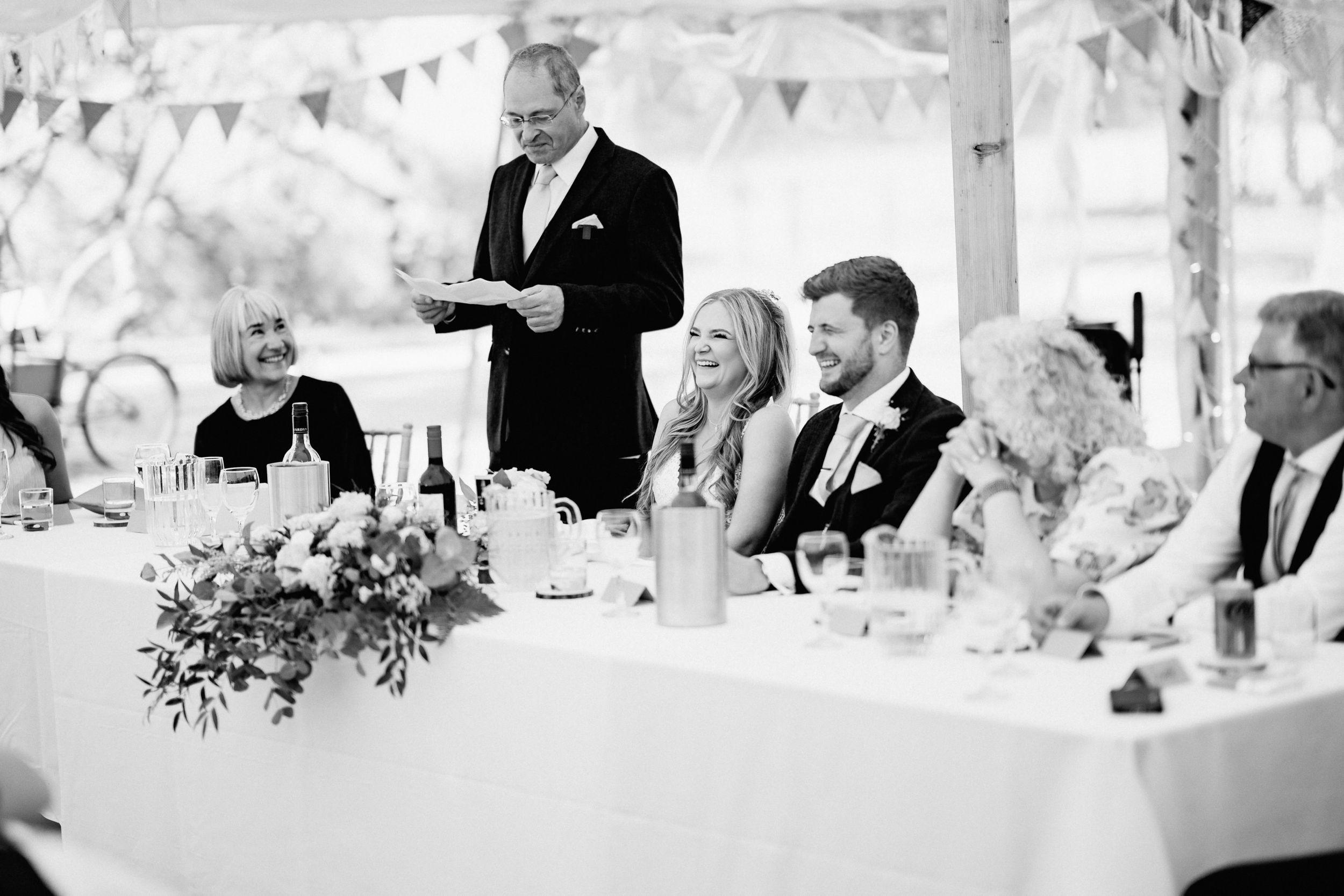 Black and white photo of a wedding reception with guests at a head table while someone delivers a speech standing.