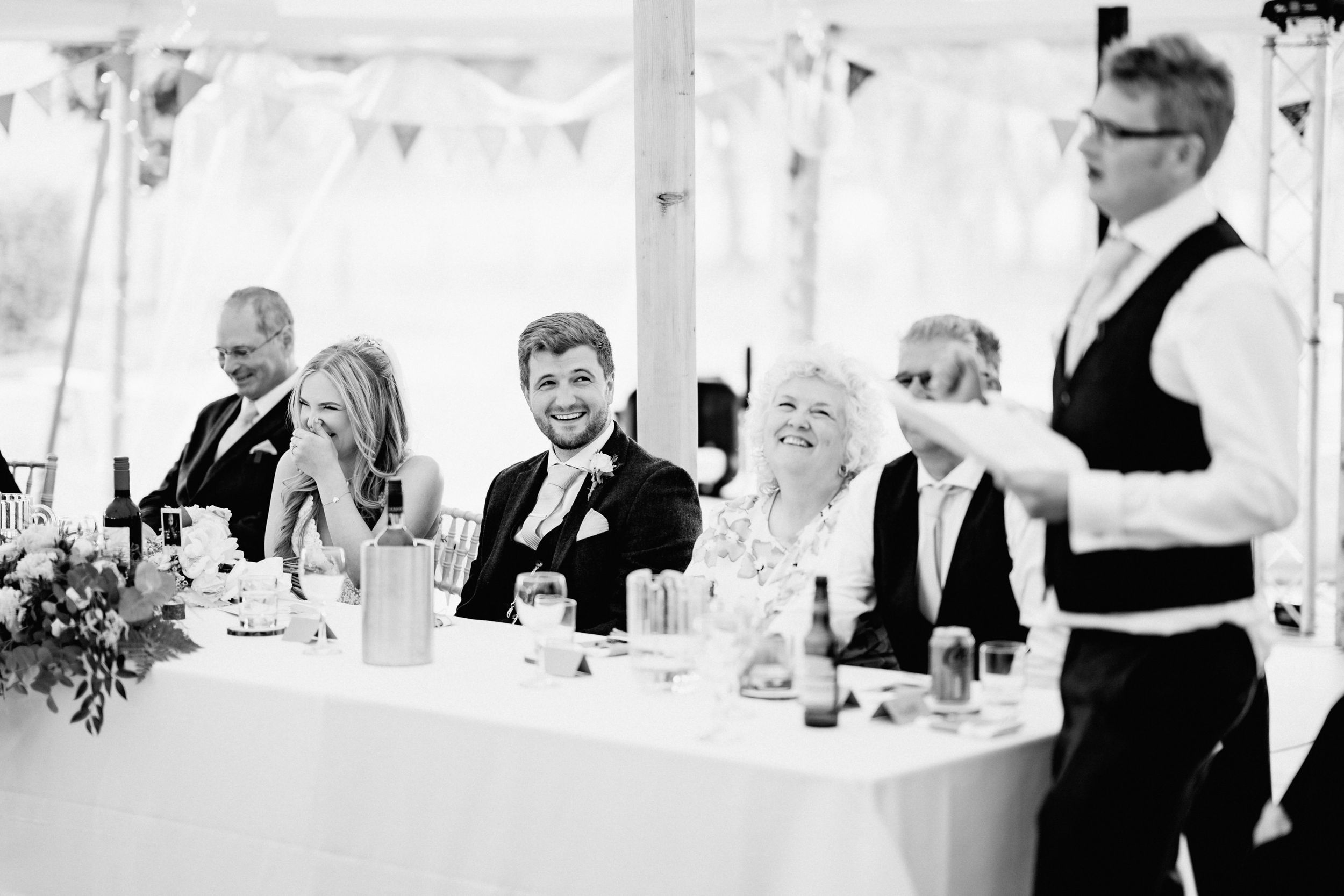 Wedding party seated at head table under white tent during reception dinner celebration.