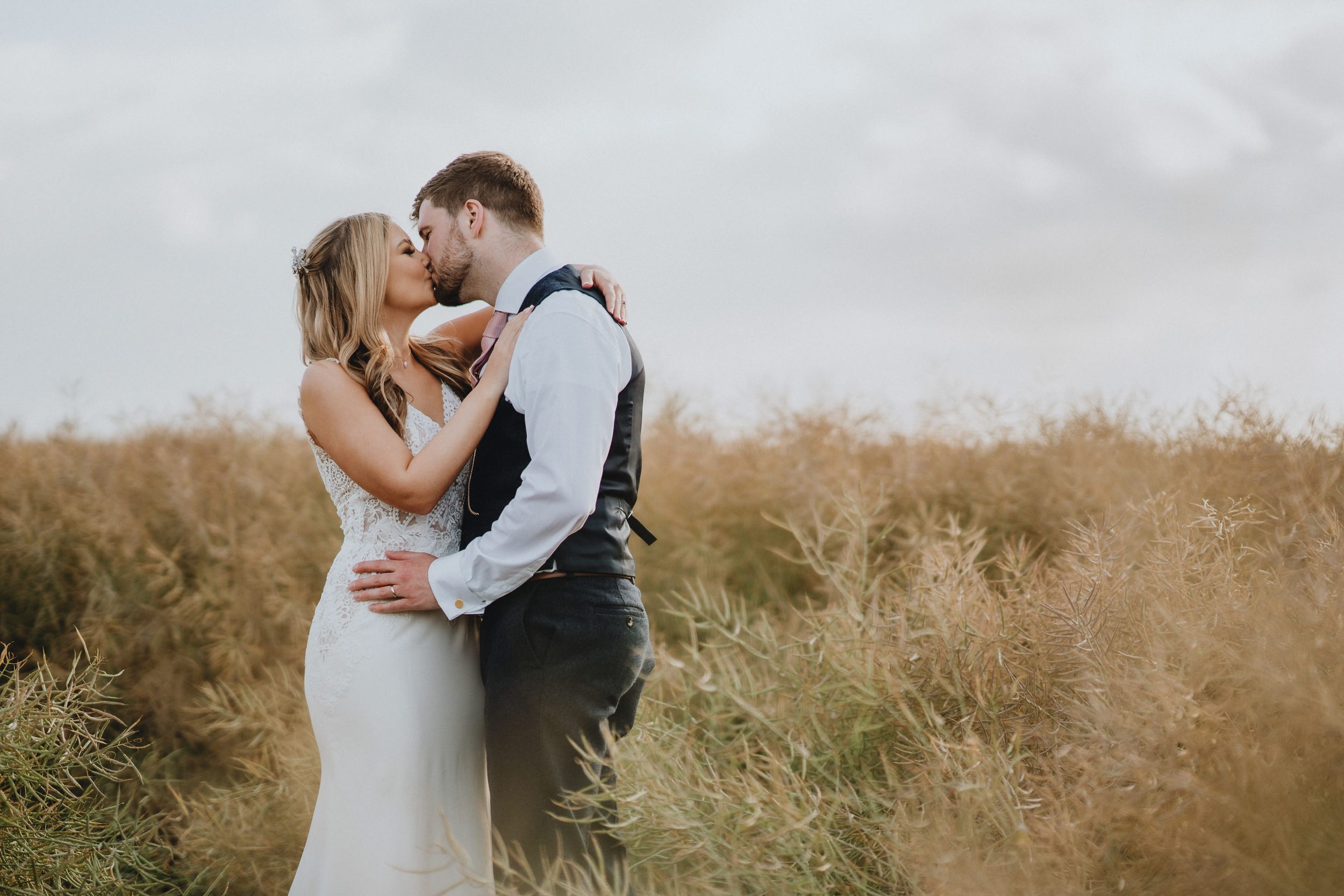 A couple embraces in a romantic kiss amidst tall golden grass on an overcast day.