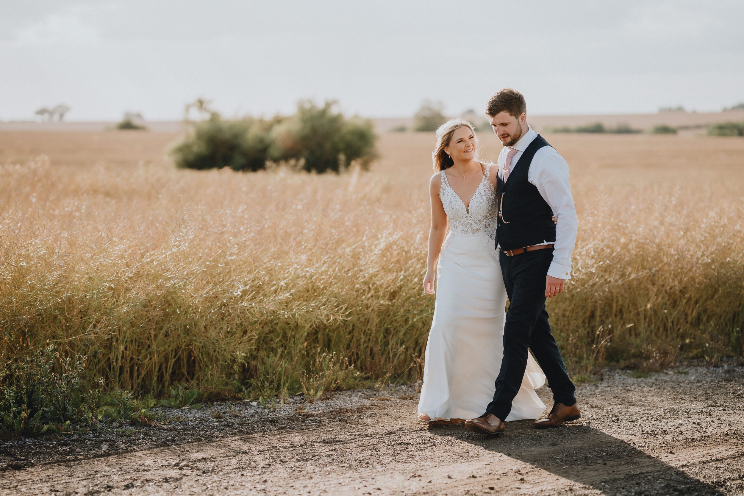 A romantic couple in wedding attire walk together on a path through a golden wheat field at sunset.