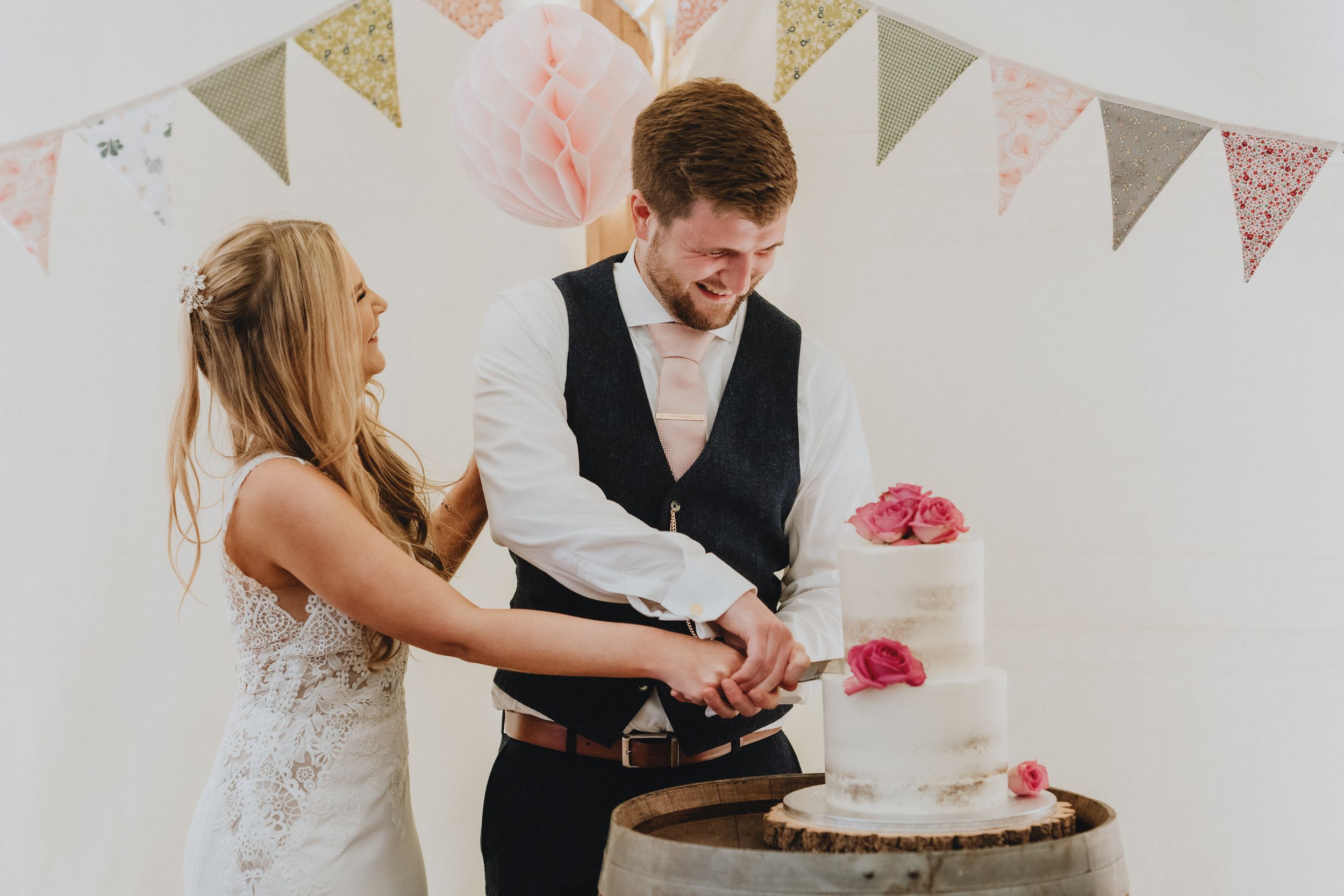 A couple cuts into their simple white wedding cake decorated with pink flowers during their reception celebration.