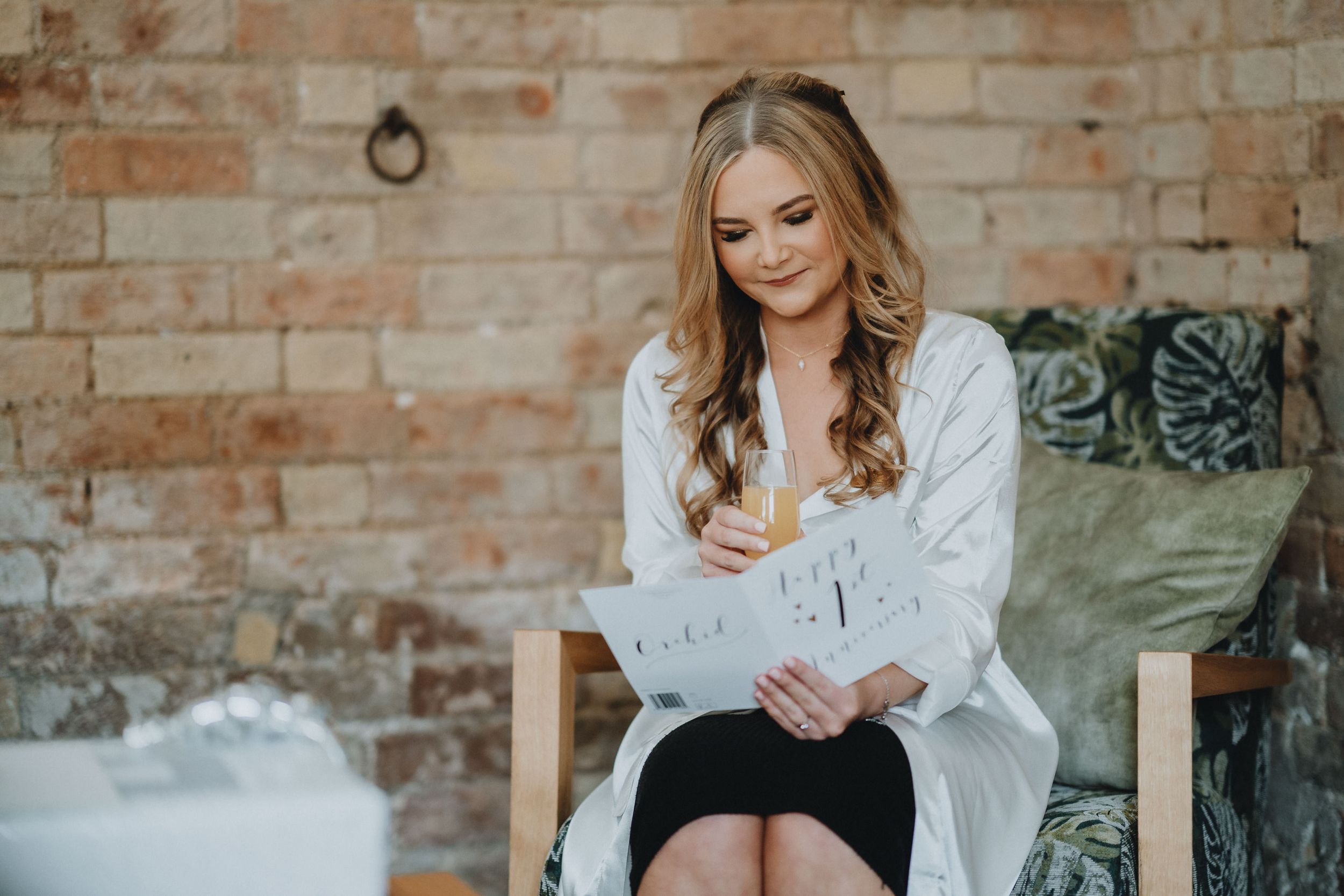 Woman in a white shirt sitting by a brick wall holding a tablet device while looking down thoughtfully.