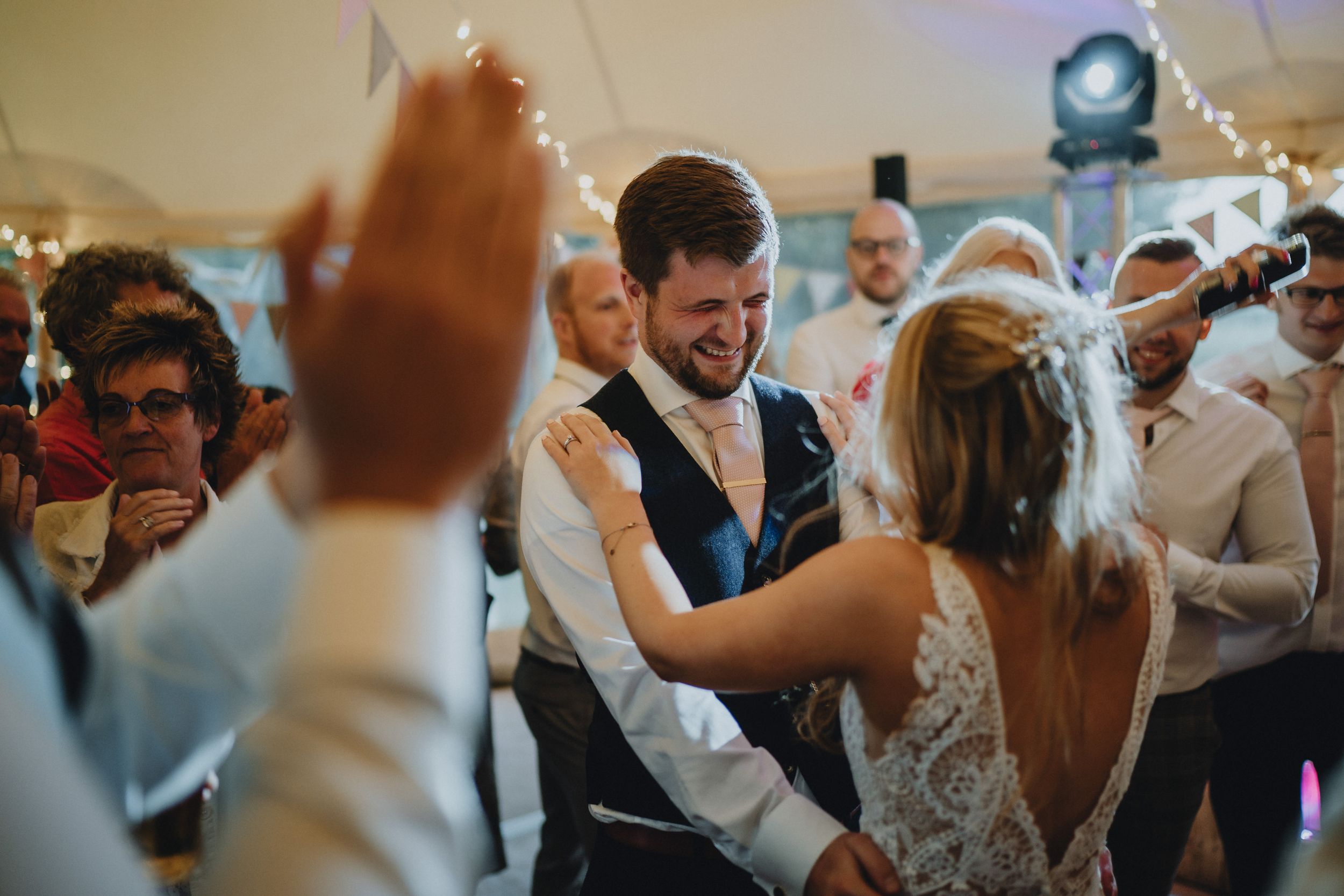 Wedding guests celebrate on a crowded dance floor during an indoor reception with atmospheric lighting.