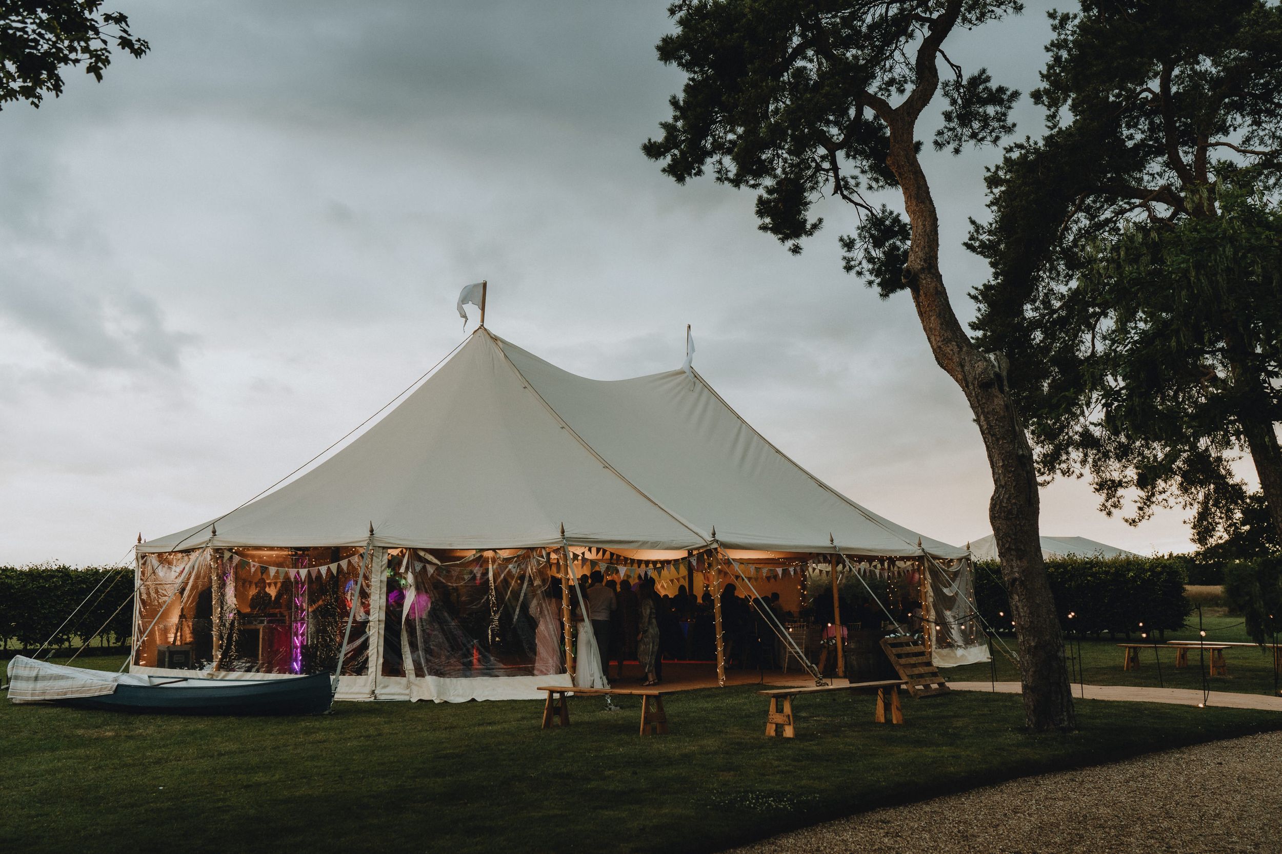 A large white sailcloth tent set up on a grass lawn with string lights and trees in the background at dusk.
