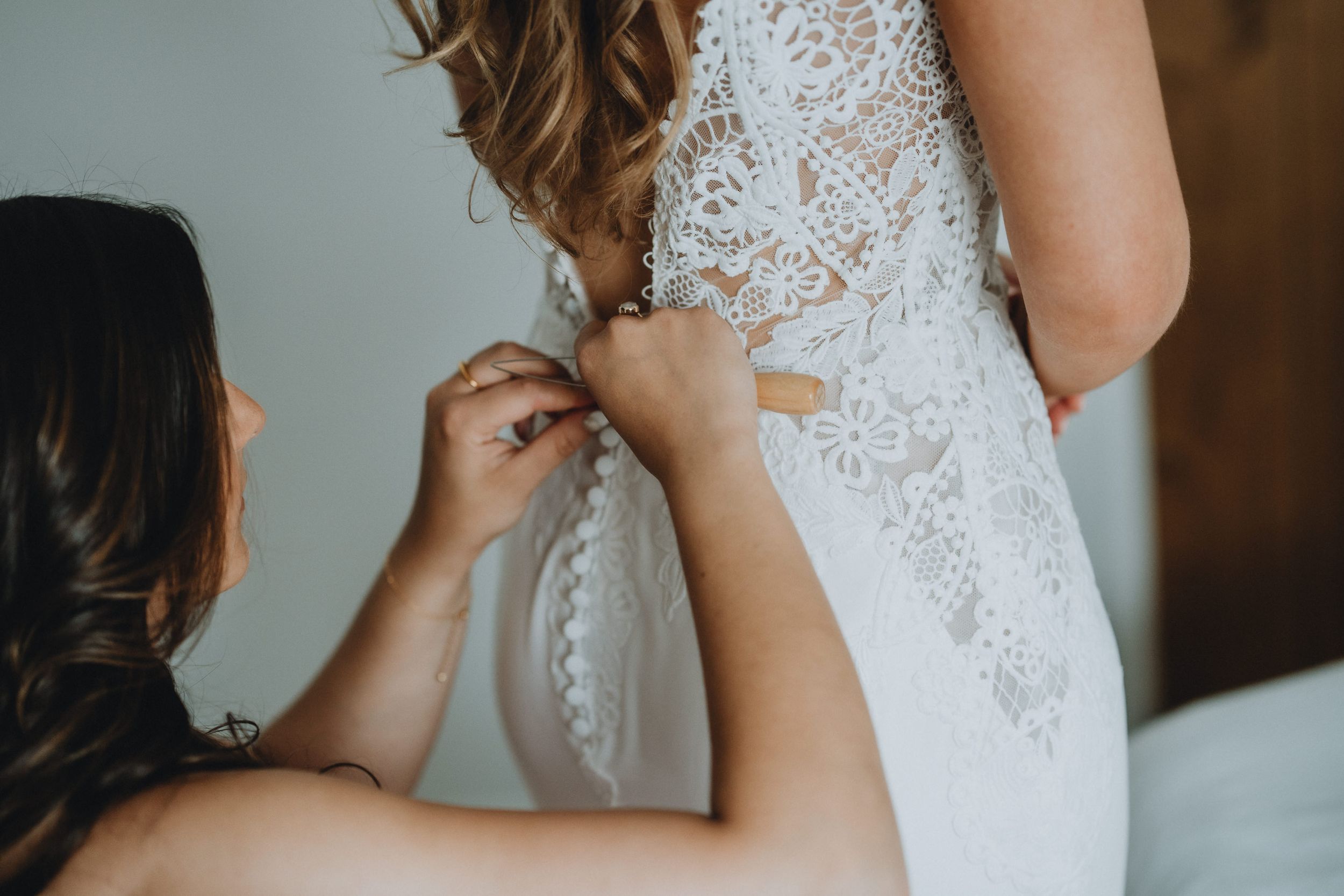 Person helping button up a delicate white lace wedding dress with intricate detailing during preparation.