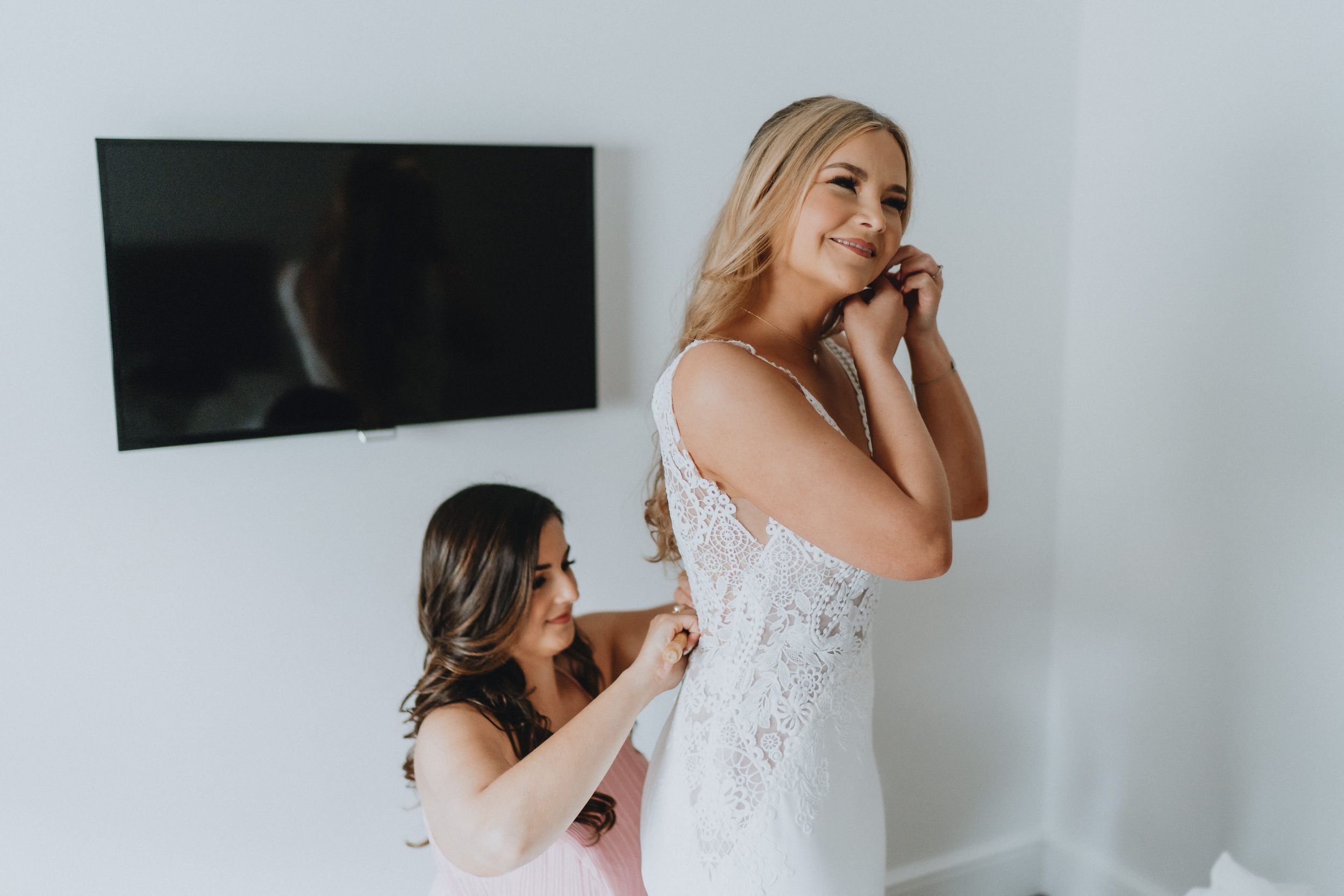 A person helps adjust a beautiful white lace wedding dress during bridal preparations in a bright room.