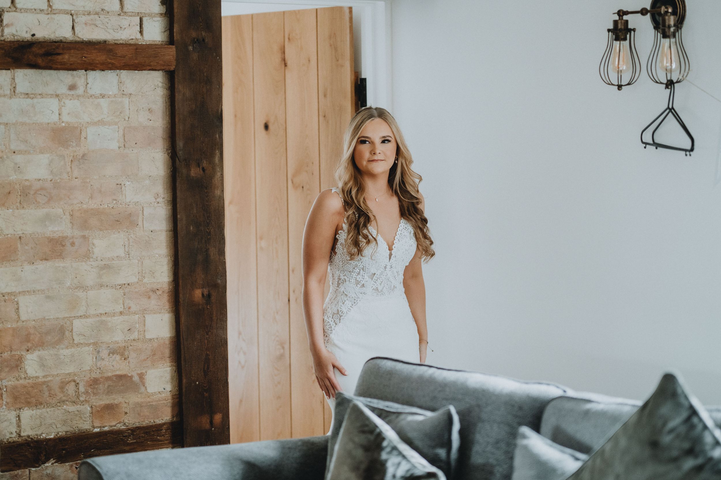 A person in a white lace wedding dress poses elegantly against exposed brick wall with industrial lighting fixtures.