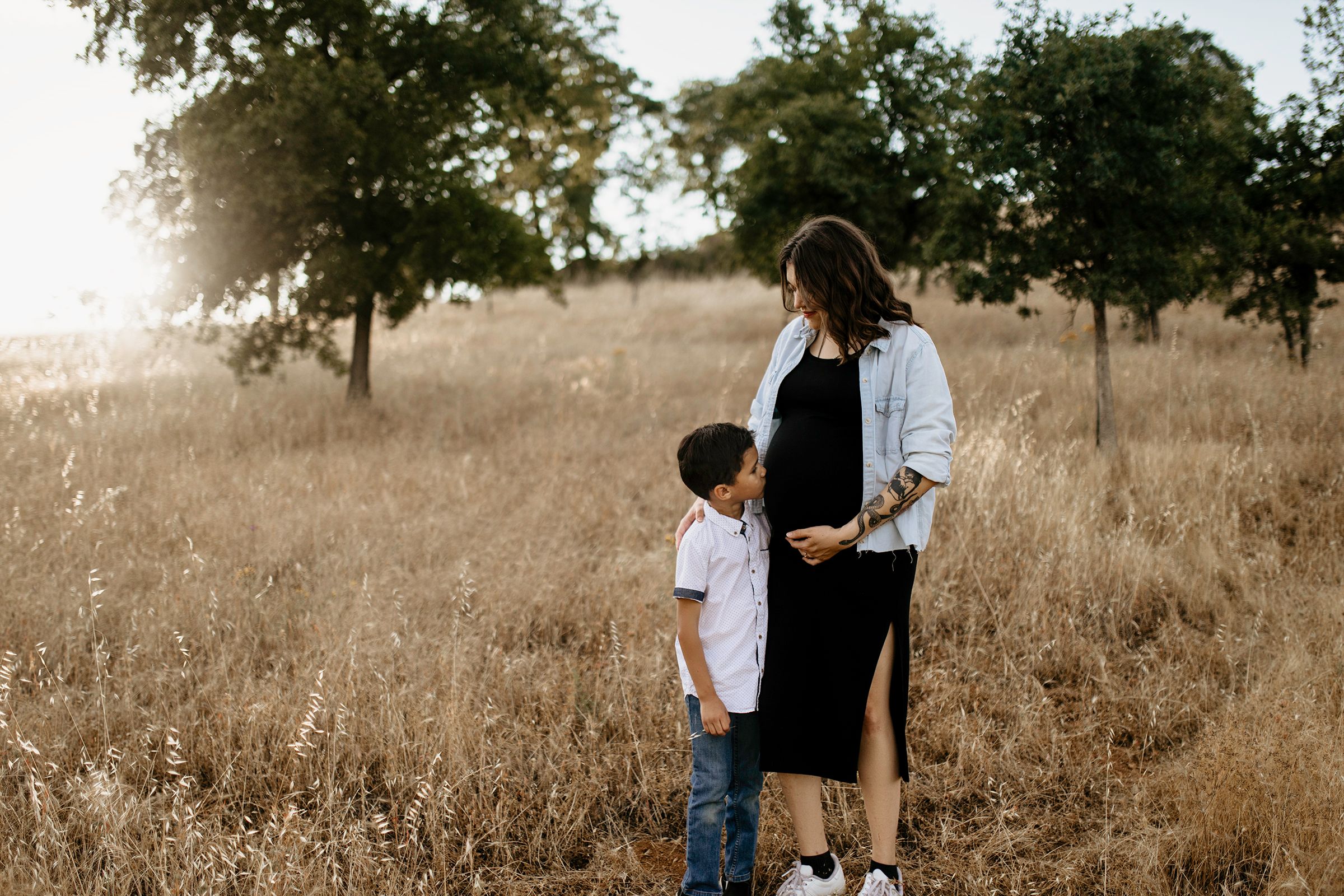 Tender family moment in golden sunlit field with tall grass and trees at sunset for maternity photos in Sacramento.