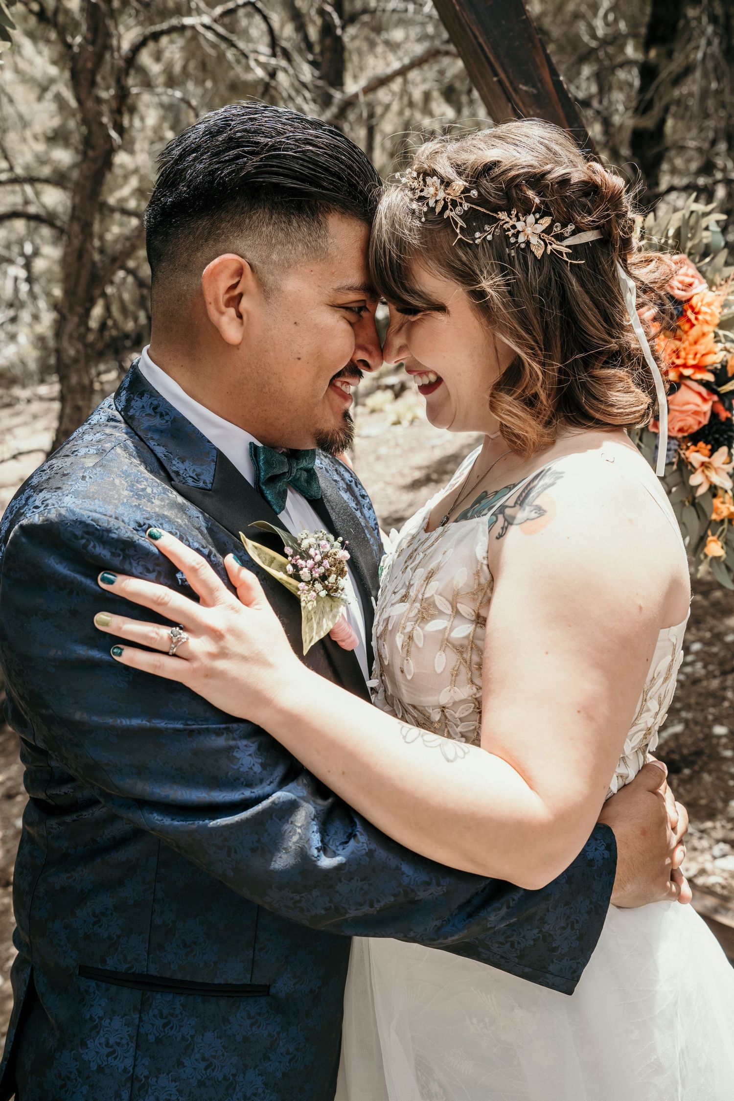 A joyful bride and groom share an intimate moment on their wedding day, smiling with foreheads touching. The groom wears a dark blue floral
