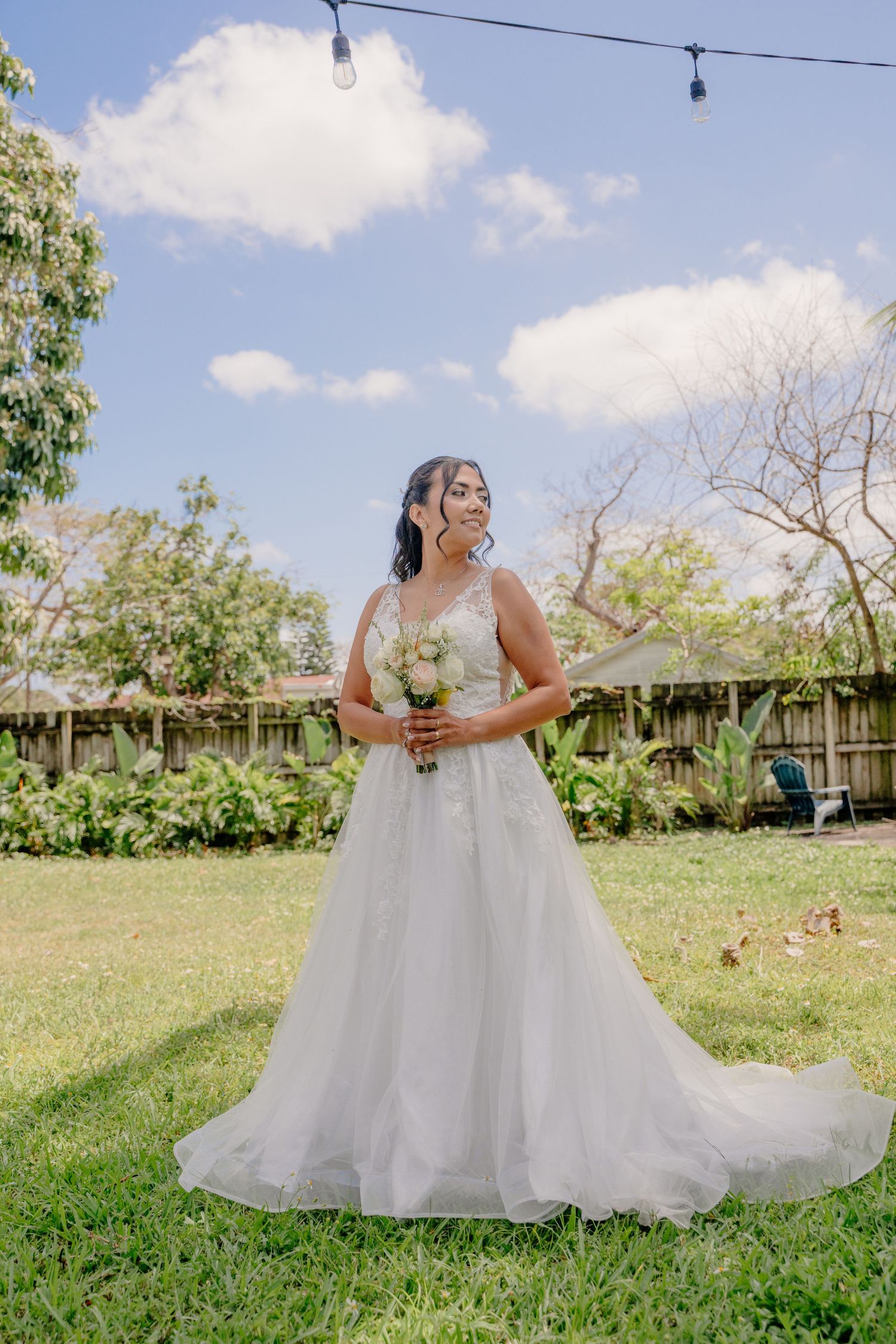 A joyful bride in a flowing white ball gown stands in a sunny garden setting holding a cream bouquet.