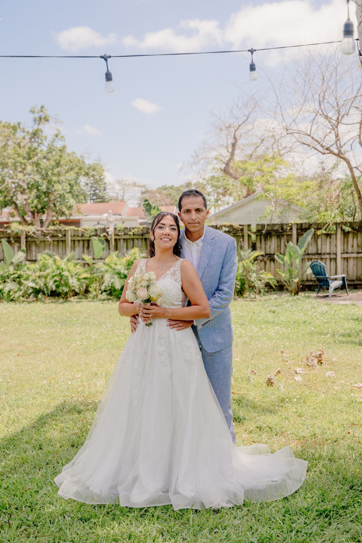 A couple embraces in a garden backyard setting with string lights overhead on their wedding day.