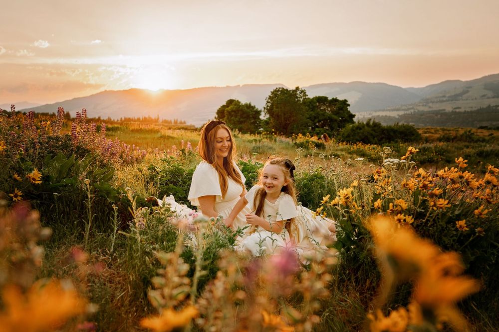 Mother and child sitting in wildflower meadow at sunset with mountains in background.