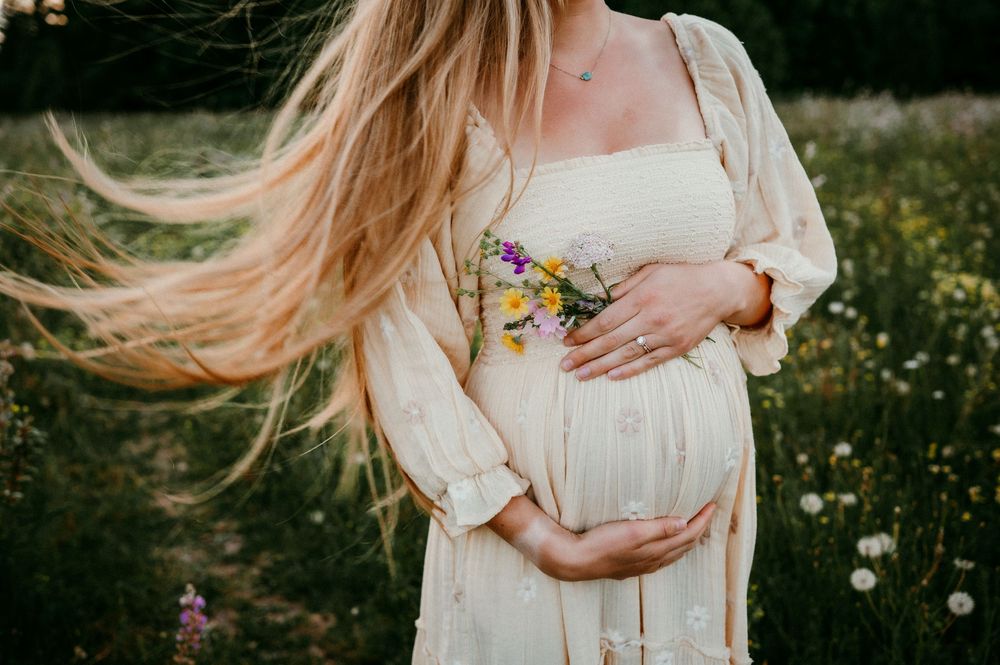 Expectant mother in white dress holds baby bump while standing in flower field.