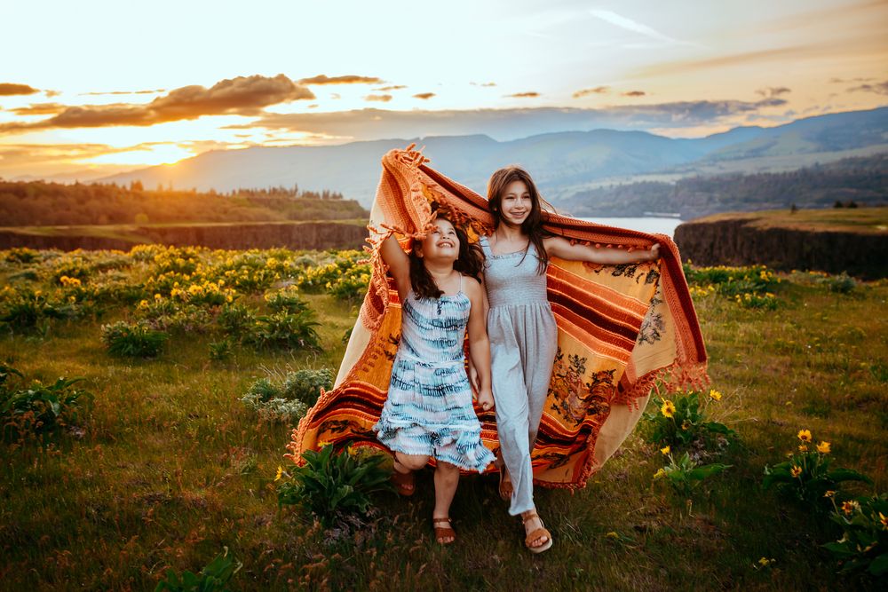 Two people share a joyful moment with a blanket in a mountain meadow at sunset.