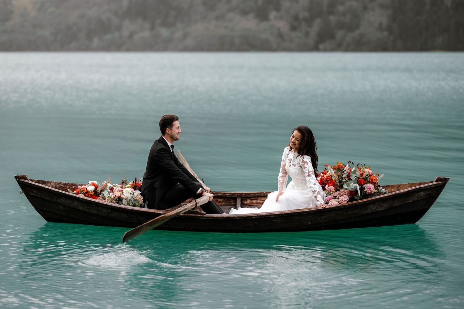 A couple sits in a wooden rowboat decorated with flowers on a tranquil turquoise lake during an intimate photo session.
