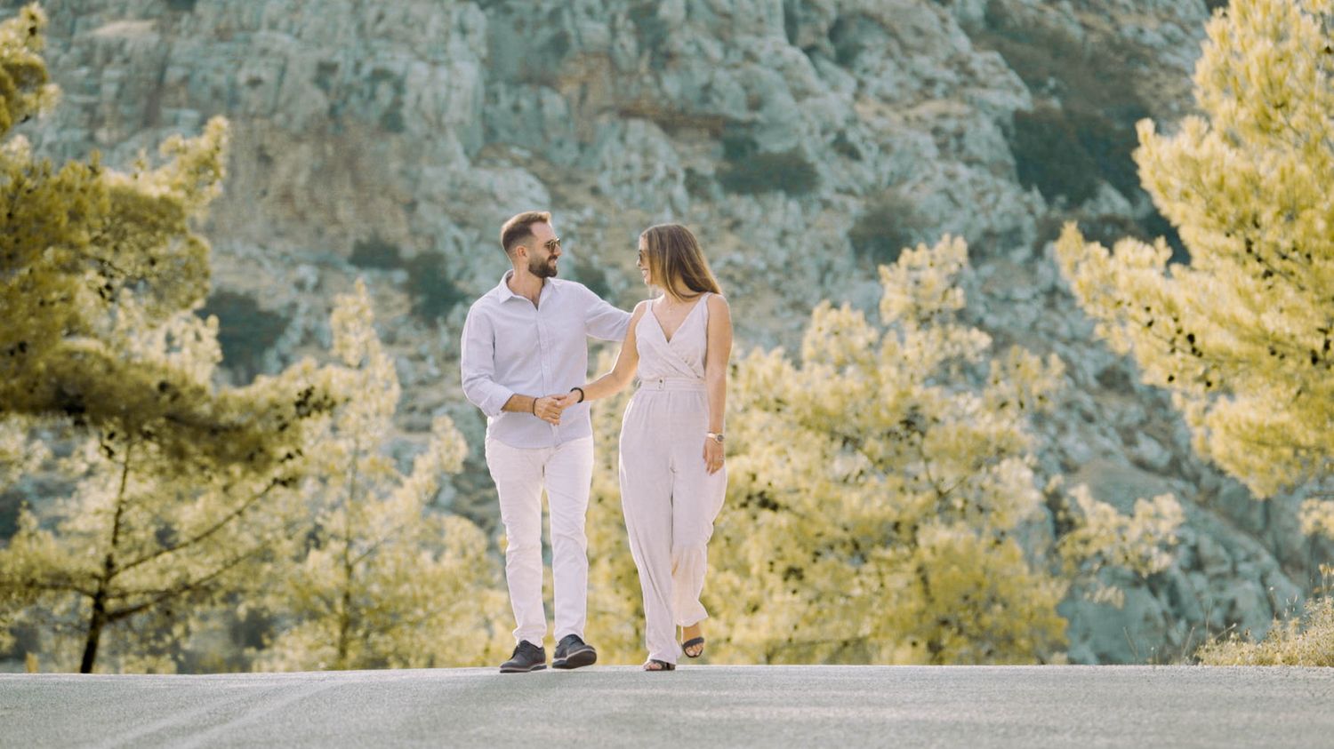A couple in white outfits walks together on a mountain path with pine trees and cliffs in the scenic background.